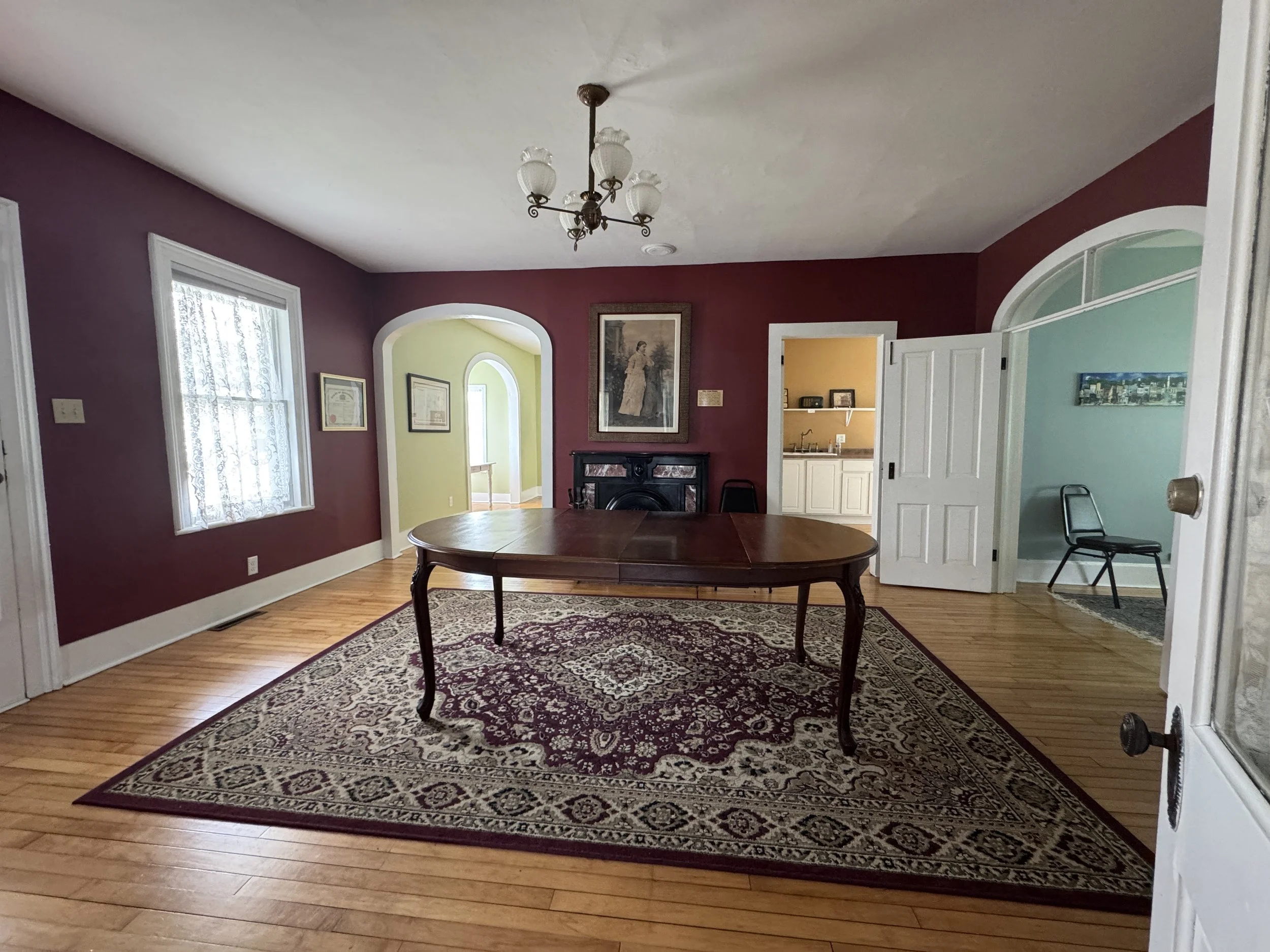 View of a dining room with a wooden table, ornate area rug, maroon walls, a chandelier, windows with lace curtains, and a doorway leading to a kitchen and other rooms, with one chair visible.
