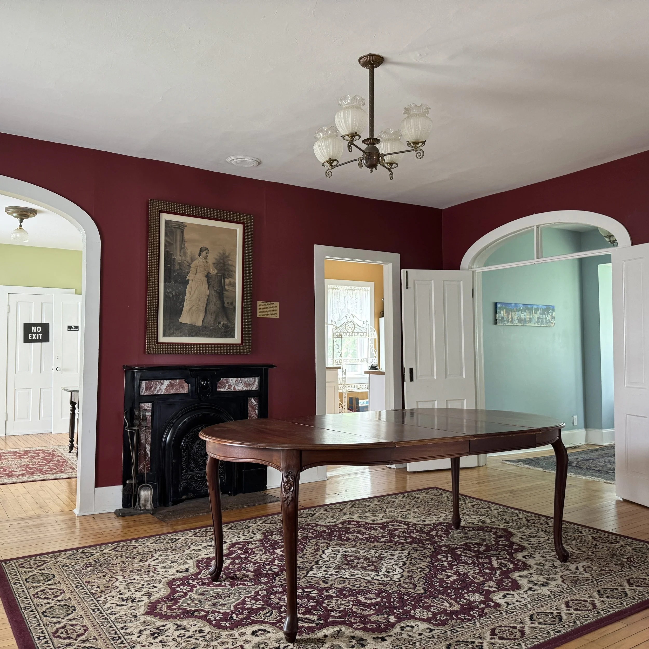Interior of a room with a wooden table on a patterned rug, red and blue painted walls, a black fireplace, and a chandelier hanging from the ceiling.
