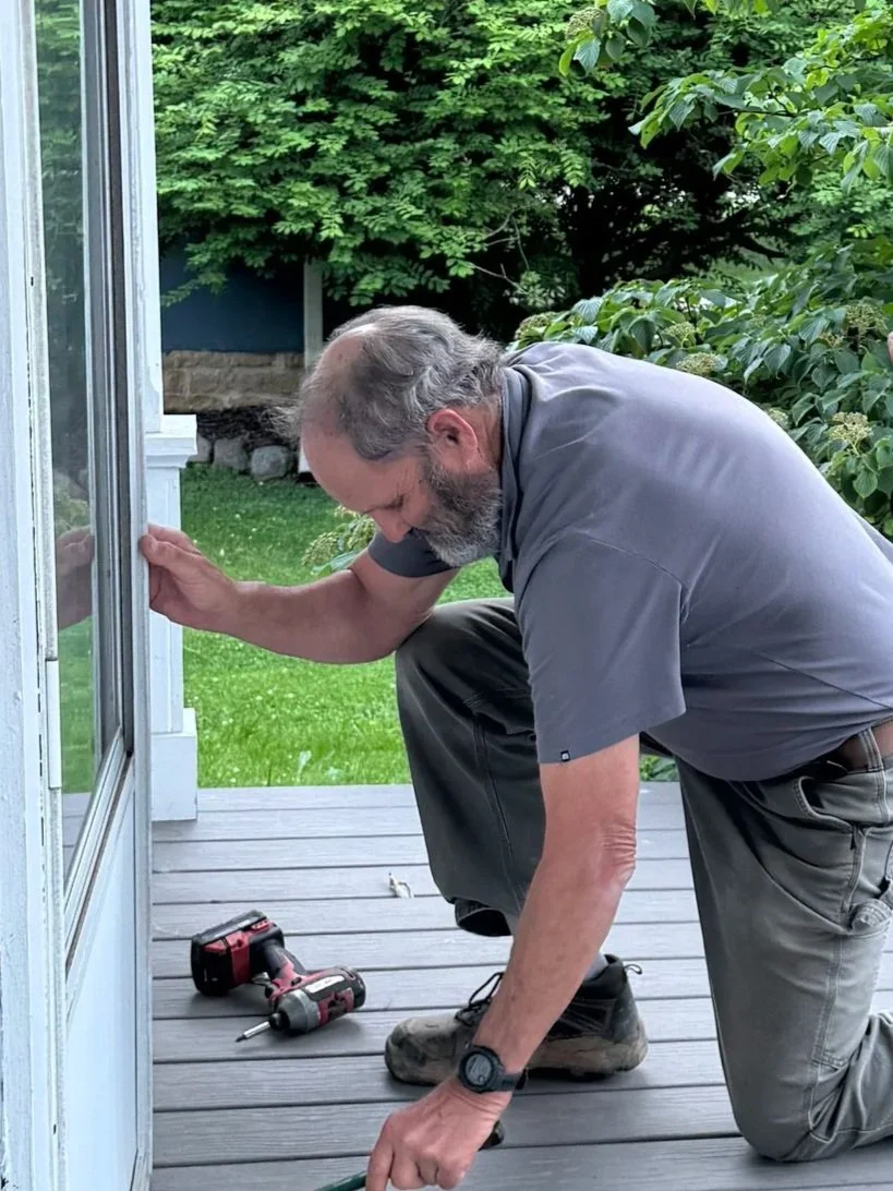 A modern house with wooden and stone exterior sits on a green hillside. Snow-capped mountains rise in the background under an overcast sky.