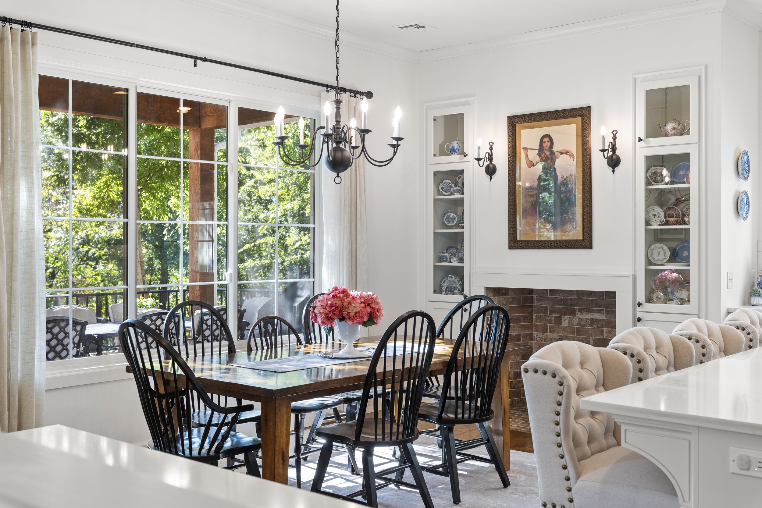 Dining room with large window, black dining chairs around a wooden table with pink flowers, white built-in shelves with decorative plates, and a chandelier.
