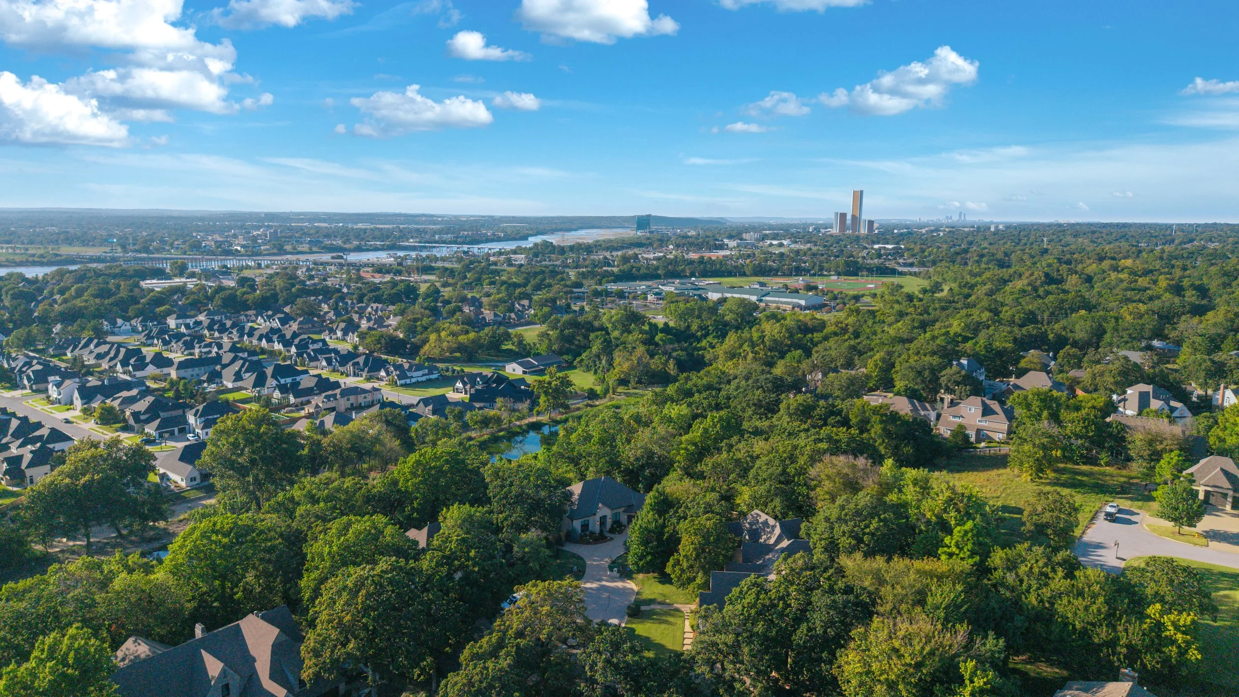 Aerial view of a suburban neighborhood with numerous houses, a small pond, lush green trees, and a distant city skyline with high-rise buildings under a blue sky with scattered clouds.
