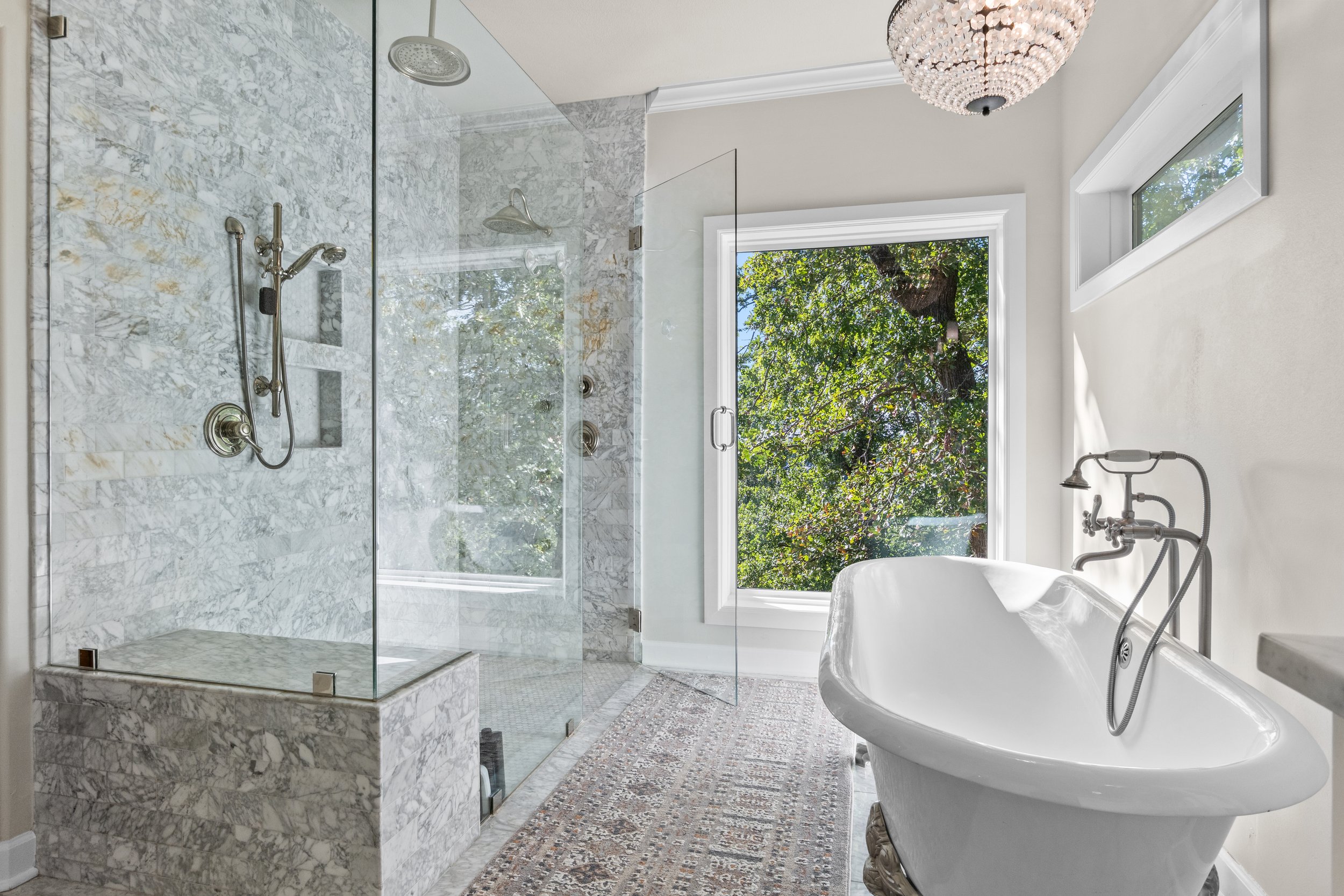 A bright bathroom with a frameless glass shower and a vintage freestanding bathtub. Large window shows green trees outside, and a decorative chandelier hangs from the ceiling.