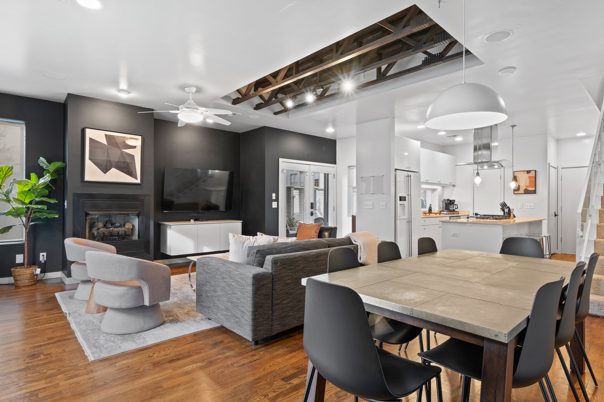 Open-concept living room and kitchen with modern decor, hardwood flooring, black accent wall, gray and white furniture, a fireplace, a wall-mounted TV, dining table with black chairs, white kitchen cabinets, island with pendant lights, and a staircas