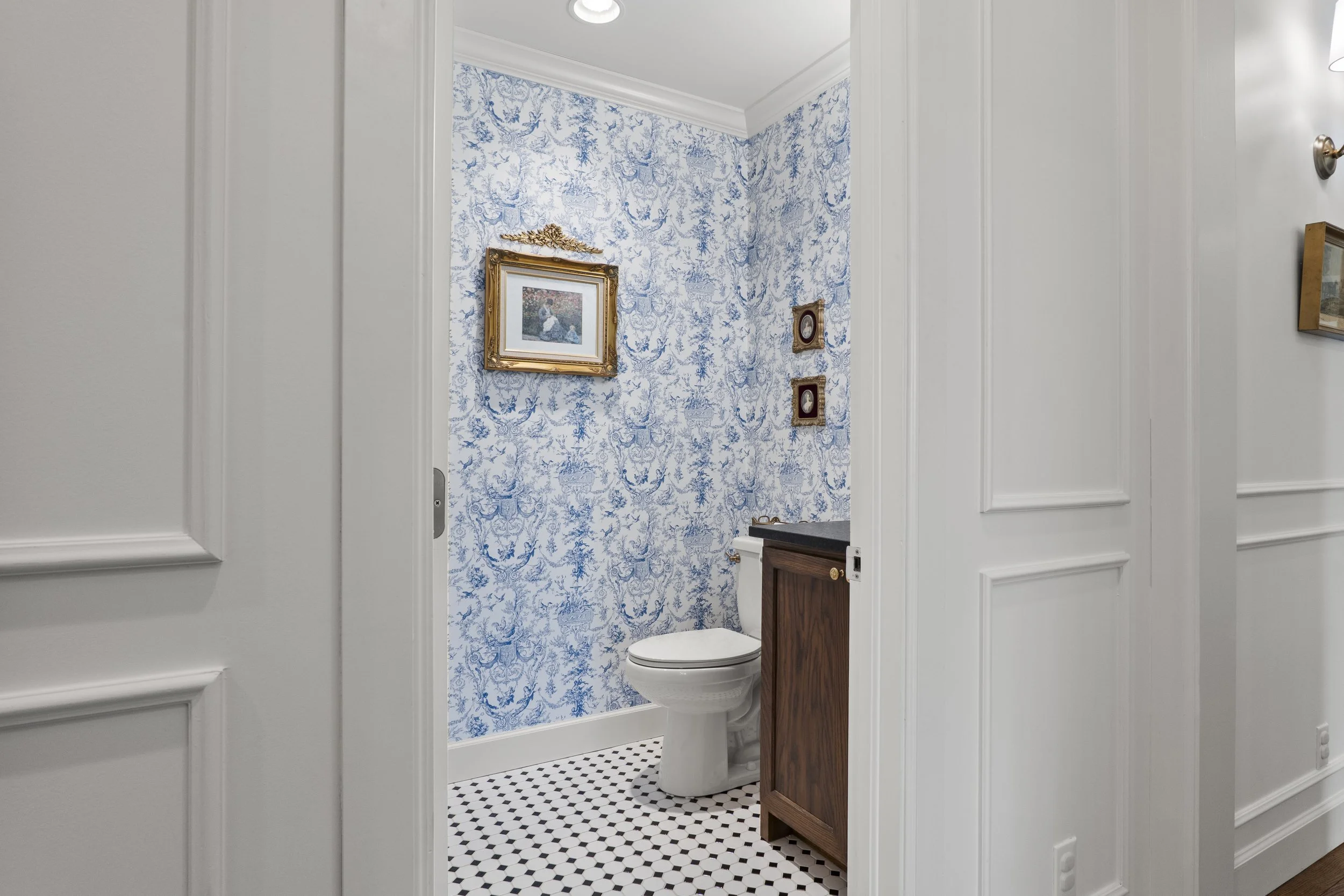 A small bathroom with blue and white patterned wallpaper, framed artwork, and two small decorative frames on the wall. The bathroom features a toilet and a wooden cabinet with a dark countertop. The floor has black and white hexagonal tiles.
