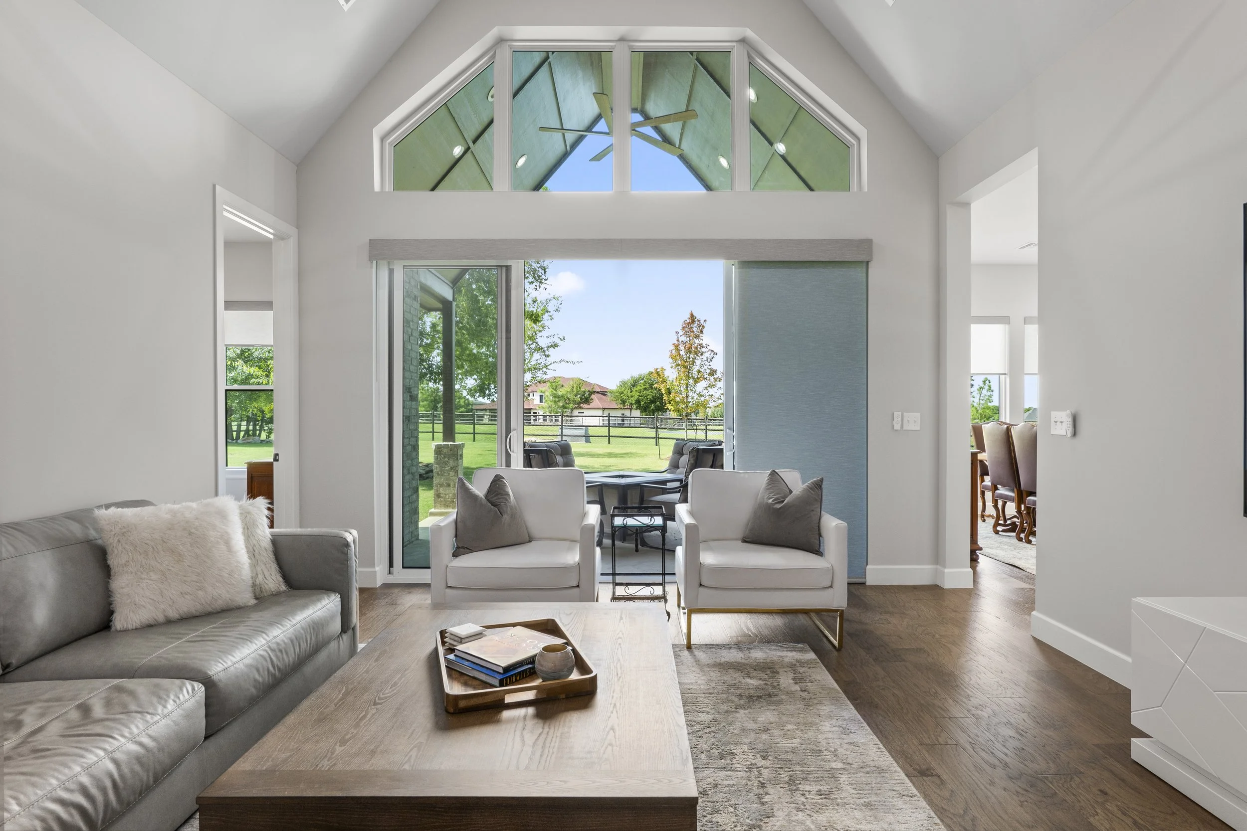 Living room with large arched window and sliding glass door leading to outdoor patio, patio with chairs, green yard, and trees in background. Interior includes gray sofa, white armchairs, wooden coffee table, and hardwood floors.