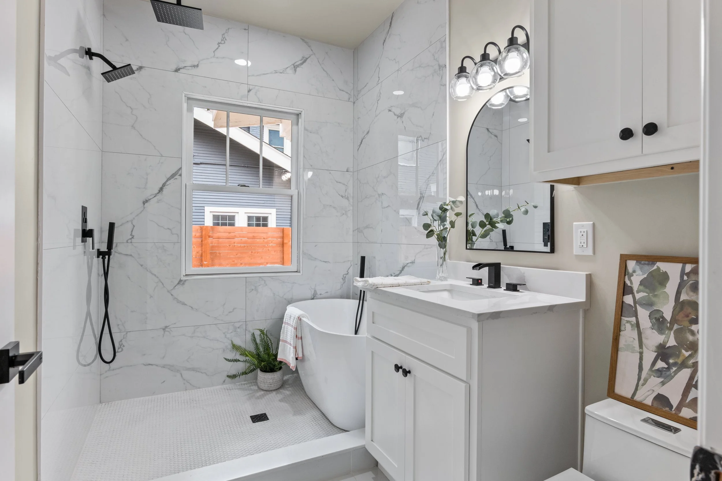 Modern bathroom with walk-in shower, freestanding bathtub, white marble walls, window, vanity with black fixtures, framed mirror, artwork, and decorative plants.