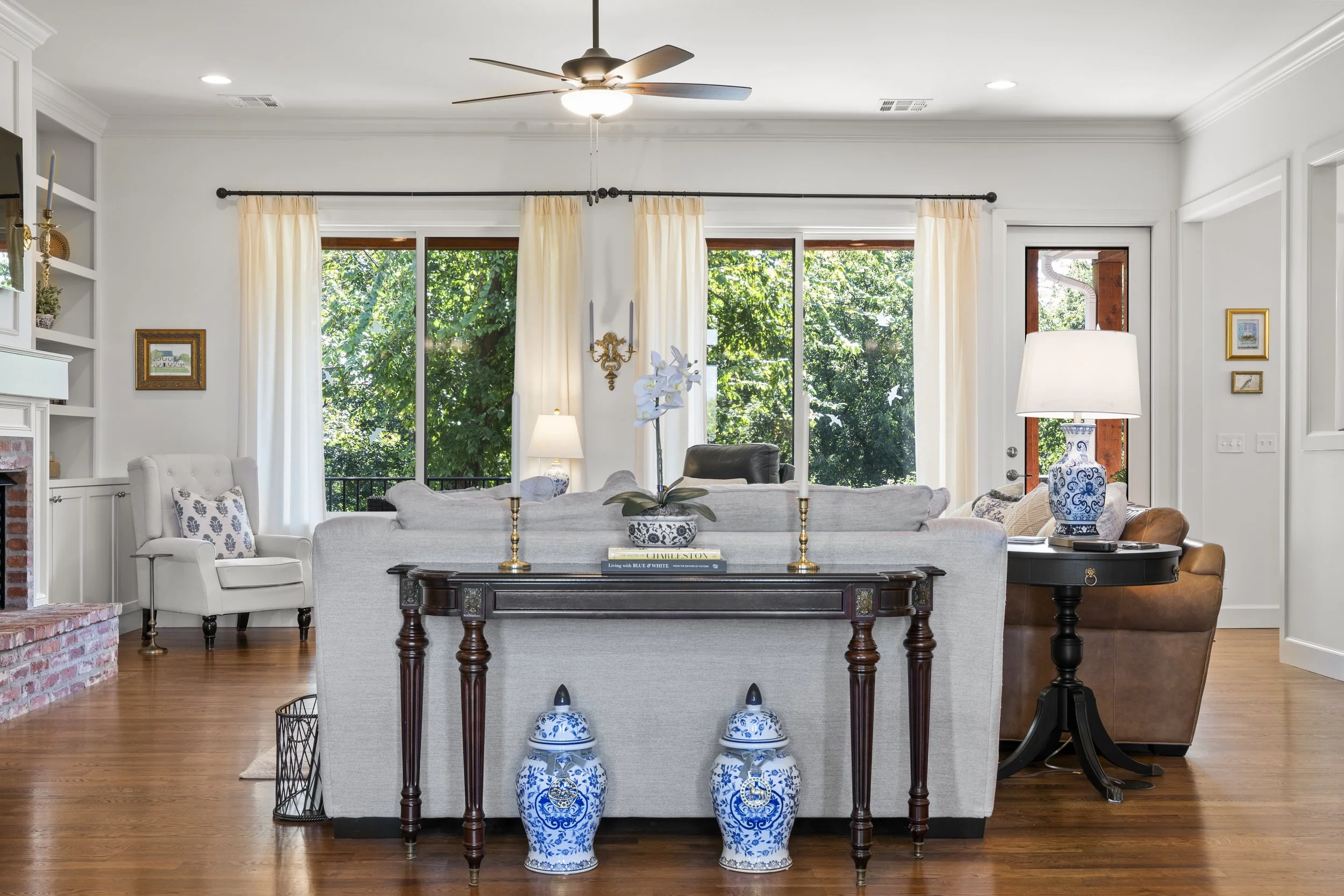 A living room with large windows showing green trees outside, furnished with a white armchair, a grey sofa, a brown leather armchair, and various lamps and decorations, including blue and white porcelain vases and candles.