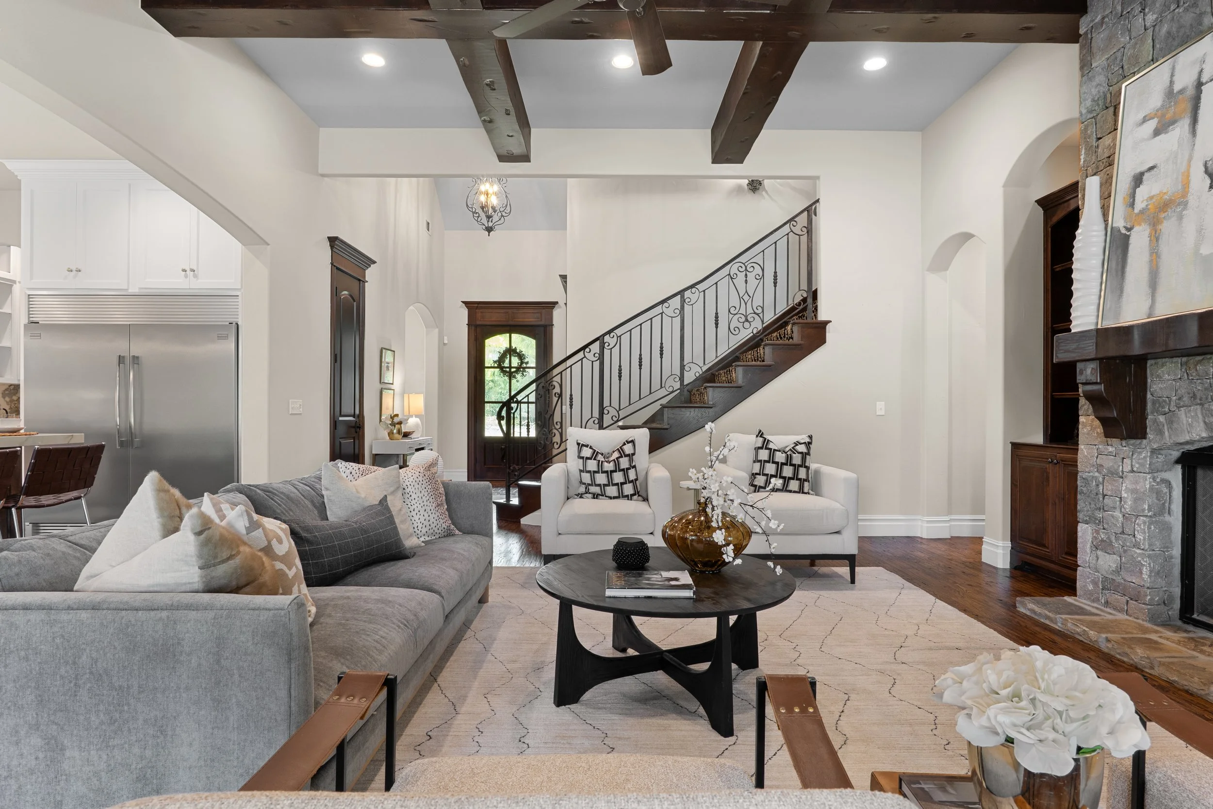 Living room with white and gray furniture, a stone fireplace, wooden ceiling beams, and a staircase with black wrought iron railing.