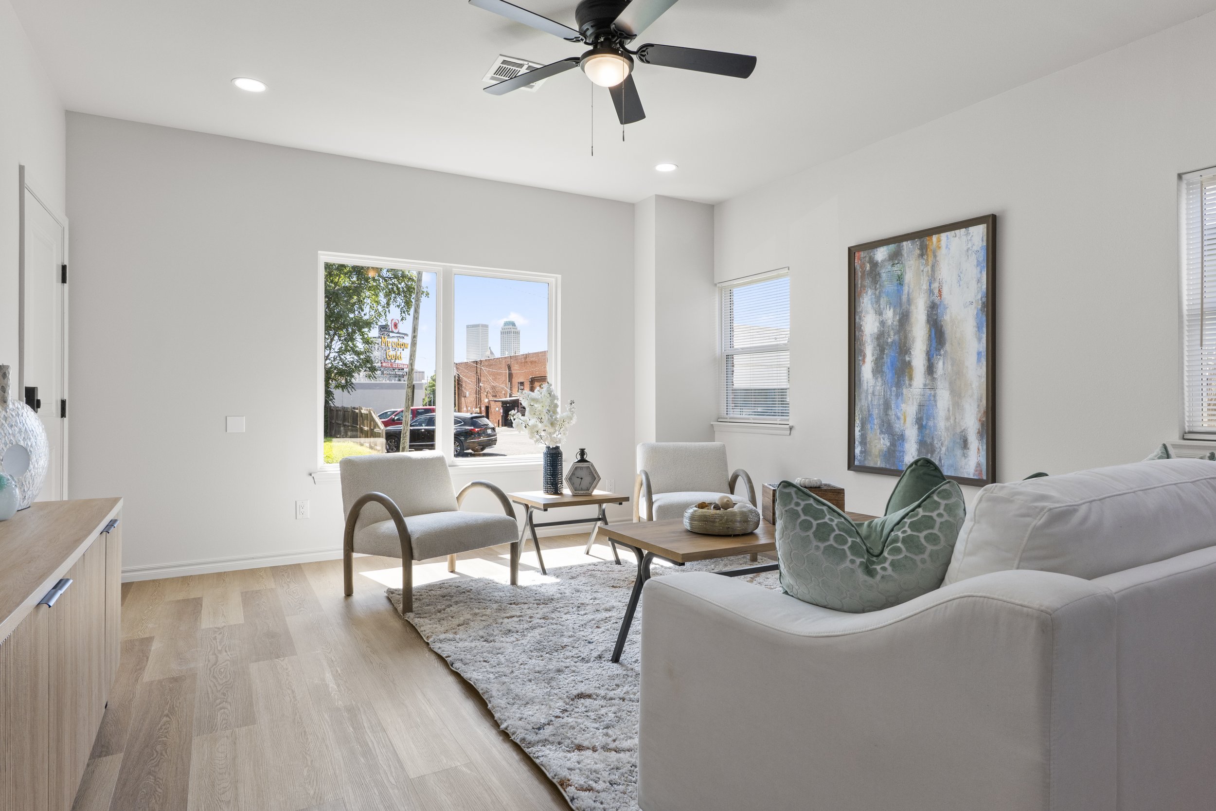 Bright living room with two beige chairs, a white sofa with green pillows, a wooden coffee table, large painting on the wall, and a window showing an outside view of parked cars and buildings.