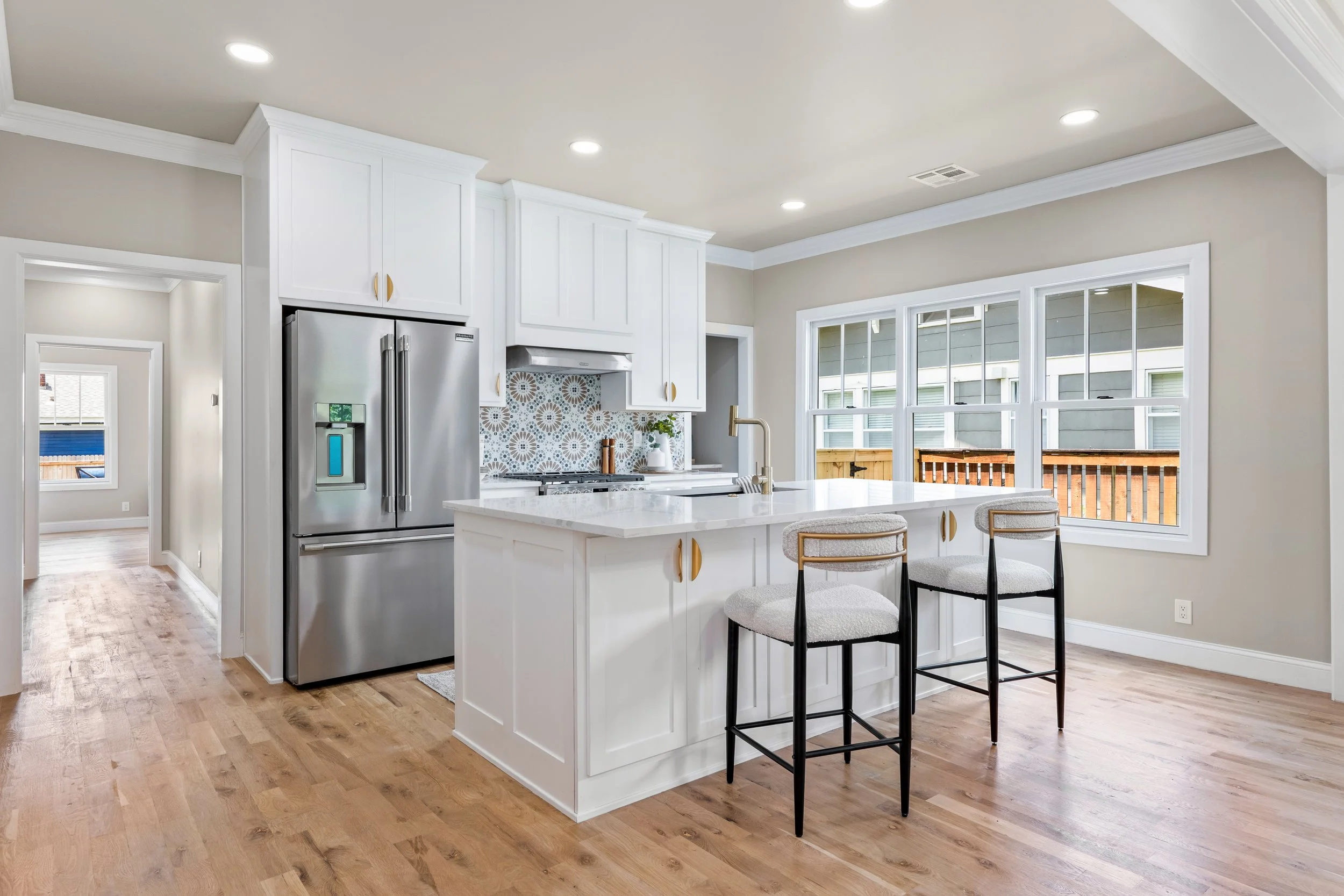 Modern kitchen with white cabinets, stainless steel refrigerator, marble countertop island with seating, large window, hardwood floors, and beige walls.