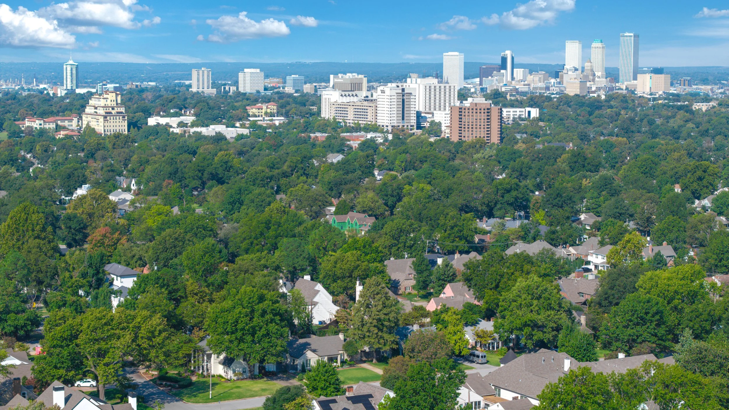 Aerial view of a city skyline with tall buildings in the distance, surrounded by a lush green suburban neighborhood with many trees and houses in the foreground.
