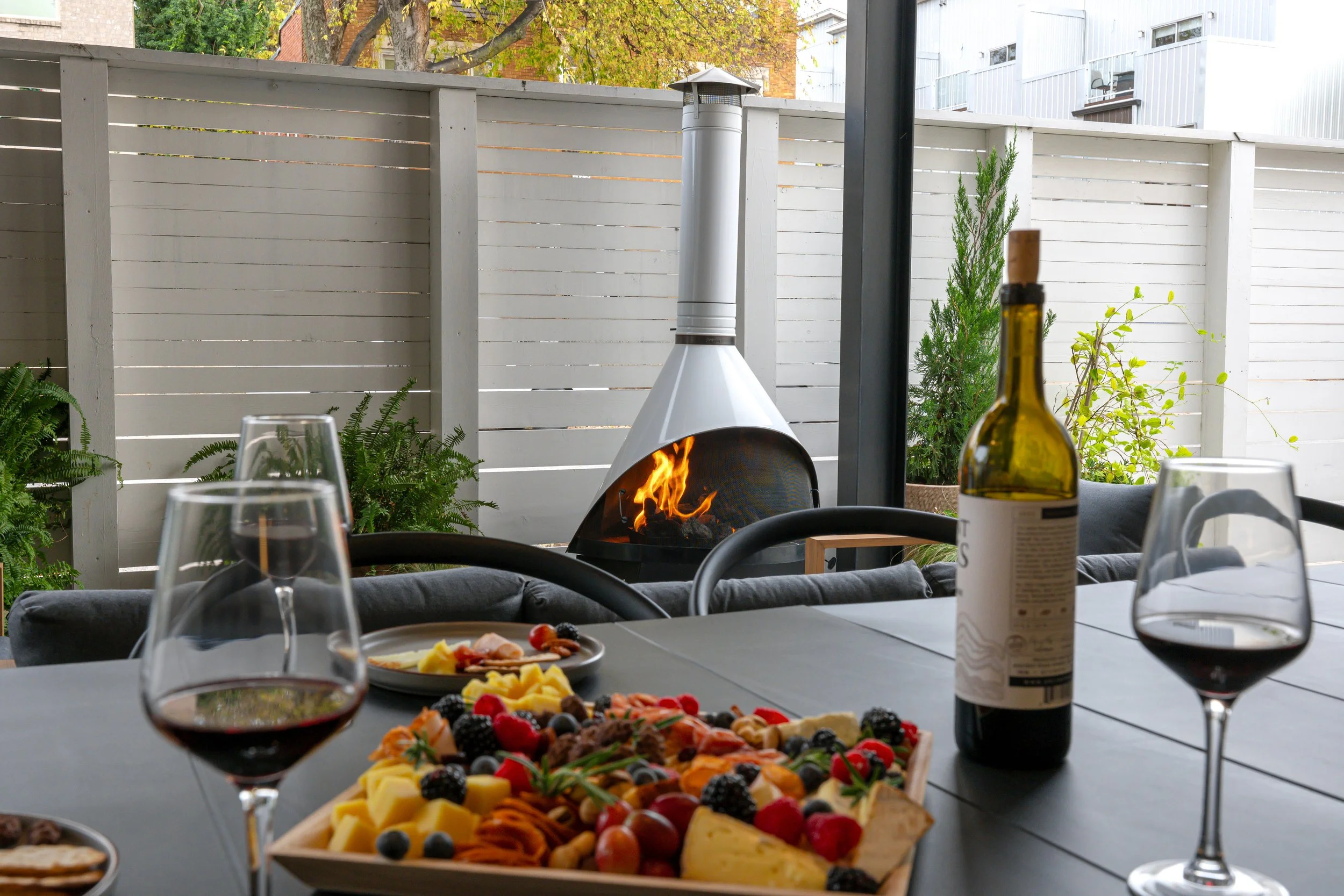 A table set for a meal with glasses of red wine, a cheese and charcuterie platter, and a bottle of wine. In the background, there is an outdoor fireplace with a flame, surrounded by a white fence and some greenery.