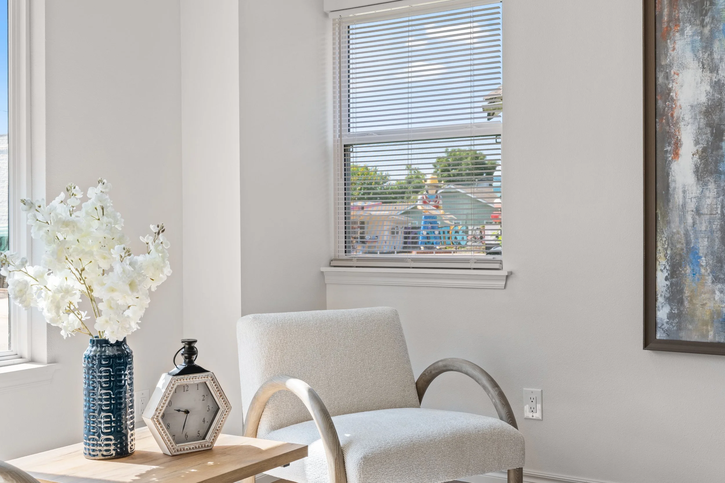 A bright living room corner with a beige upholstered armchair, a wooden side table holding a white clock and a blue vase with white flowers, and a window with white blinds showing a playground outside.