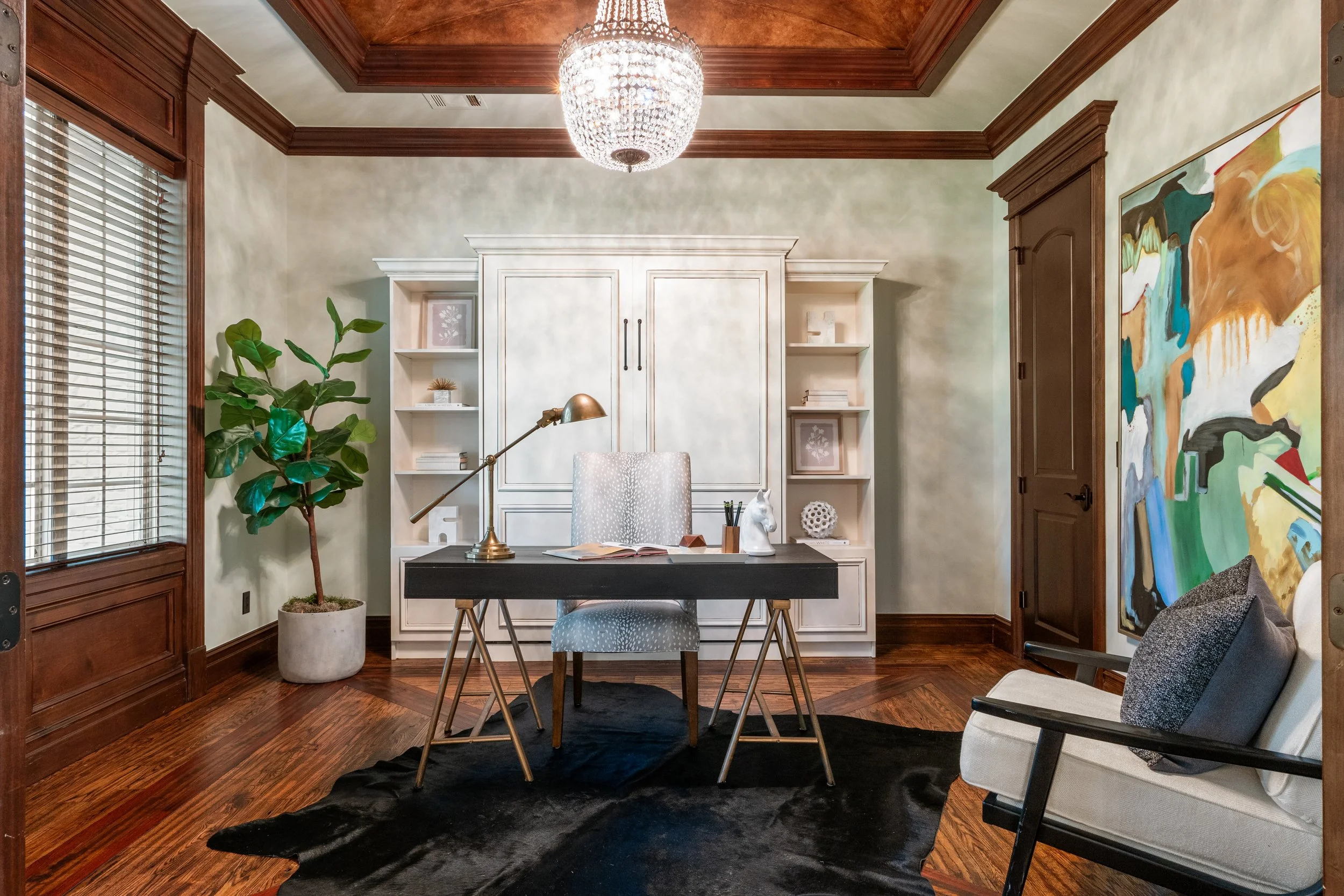 Home office with white desk, grey chair, black rug on hardwood floor, large potted plant, abstract painting, bookshelf, wooden door, and chandelier