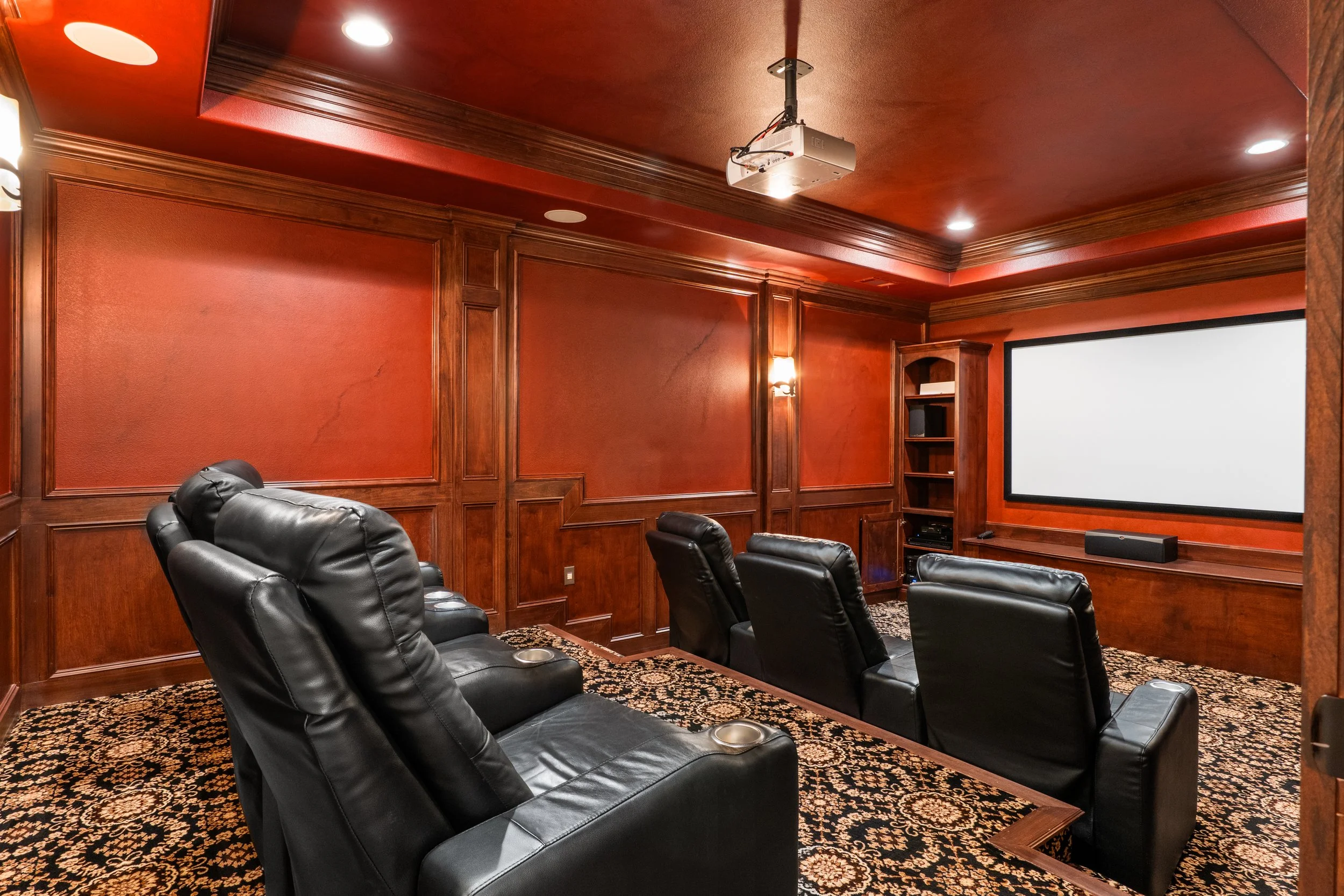 A home theater room with six black leather chairs facing a large white screen. The room has warm wood-paneled walls, ornate crown molding, and a patterned carpet. A ceiling-mounted projector is visible above, and a soundbar is below the screen.