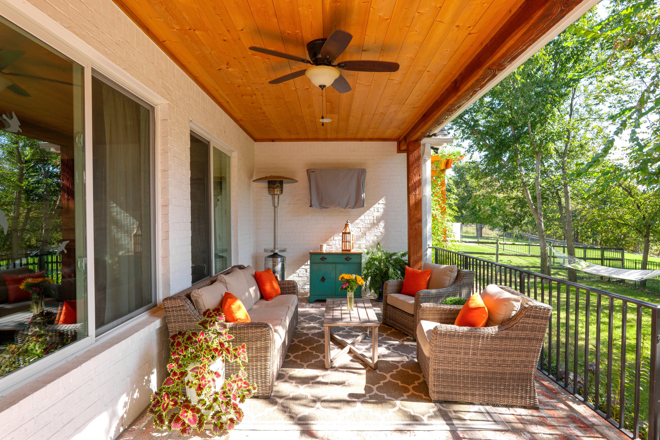 A cozy outdoor porch with wicker furniture, orange pillows, a wooden table with a flower vase, a large potted plant, a ceiling fan, and a view of trees and a grassy yard.