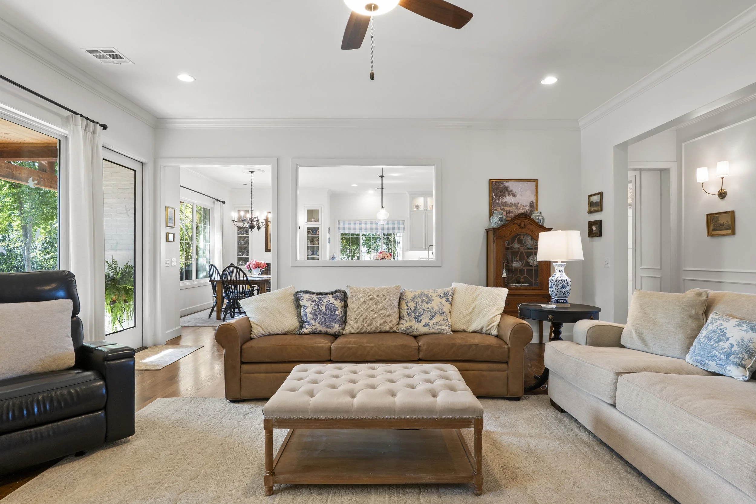 Living room with beige and black couches, wooden coffee table, and a view into the dining area, living room wall with small paintings, window with curtains, and a ceiling fan.