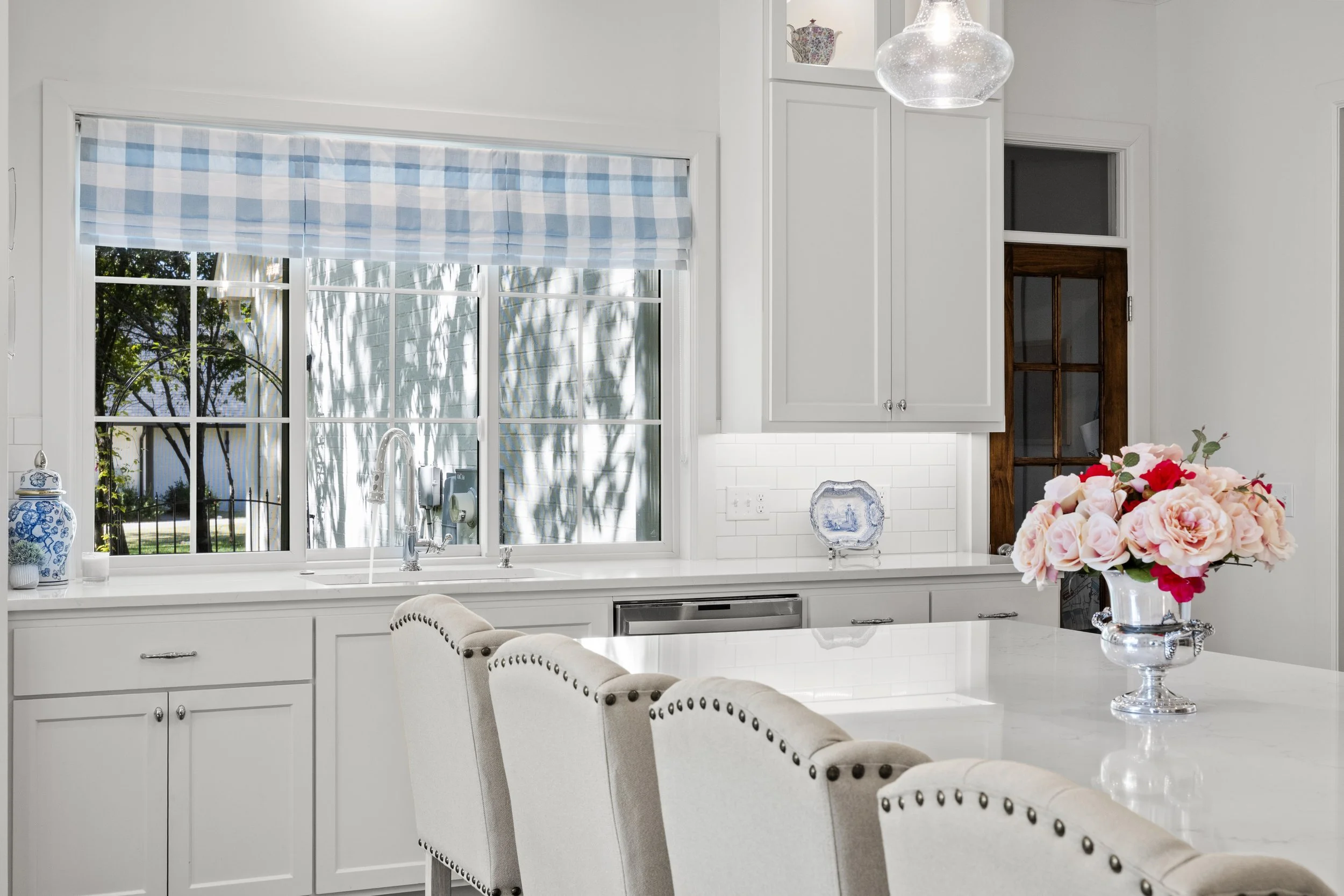 Bright kitchen with white cabinets, a window with a blue and white checkered valance, a kitchen island with beige upholstered chairs, and a vase of pink and red flowers on the counter.