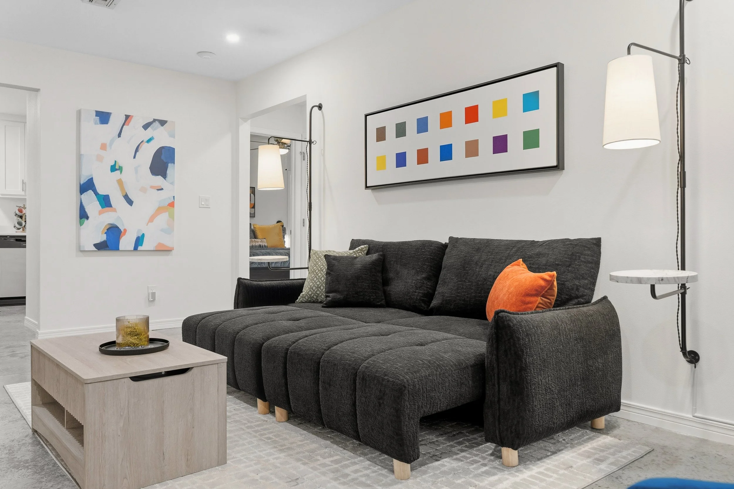 Living room with black sofa, orange and patterned pillows, abstract artwork with colorful squares, wall-mounted lamps, light-colored coffee table, white walls, part of kitchen in background.