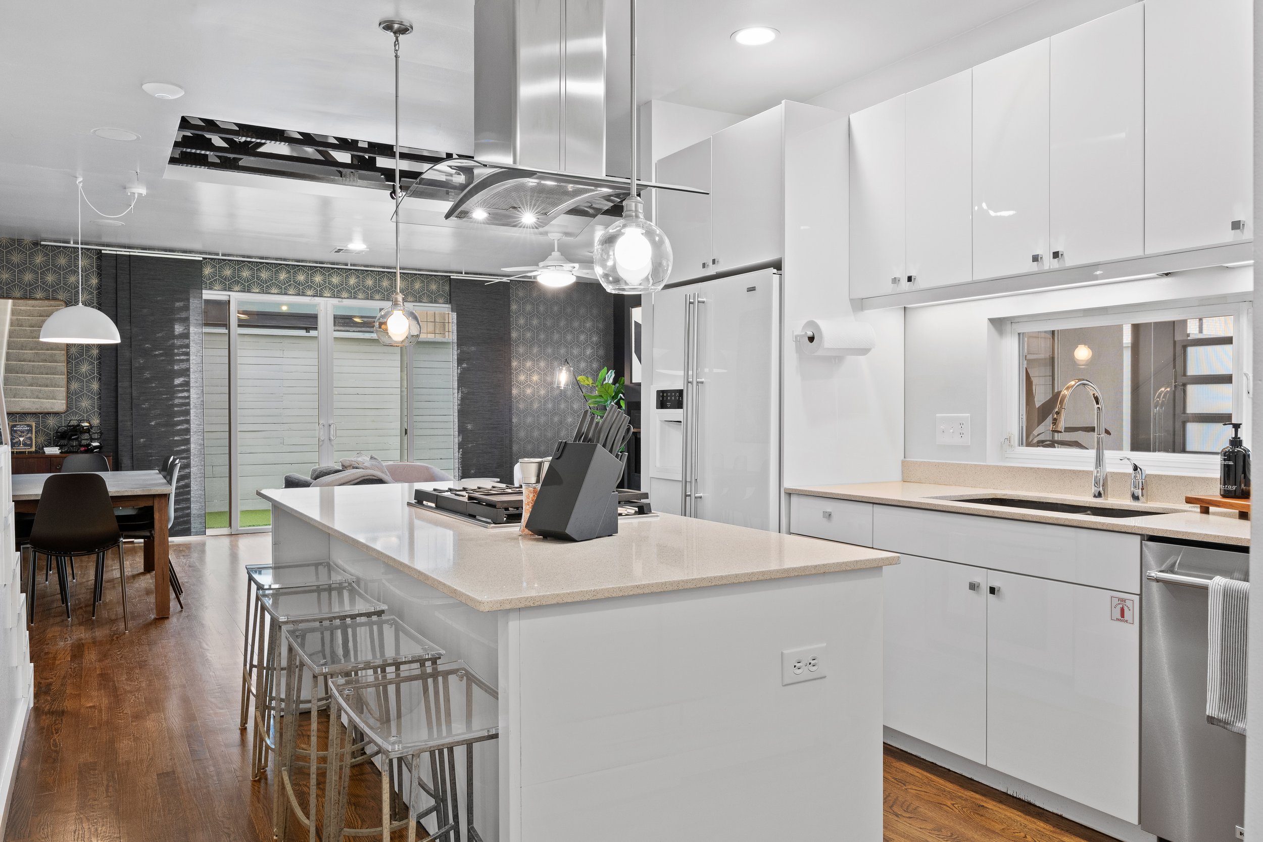 Modern kitchen with white cabinets, beige countertop, stainless steel appliances, and a kitchen island with barstools. In the background, a dining area with a wooden table and black chairs, and sliding glass doors leading outside. Wooden flooring thr
