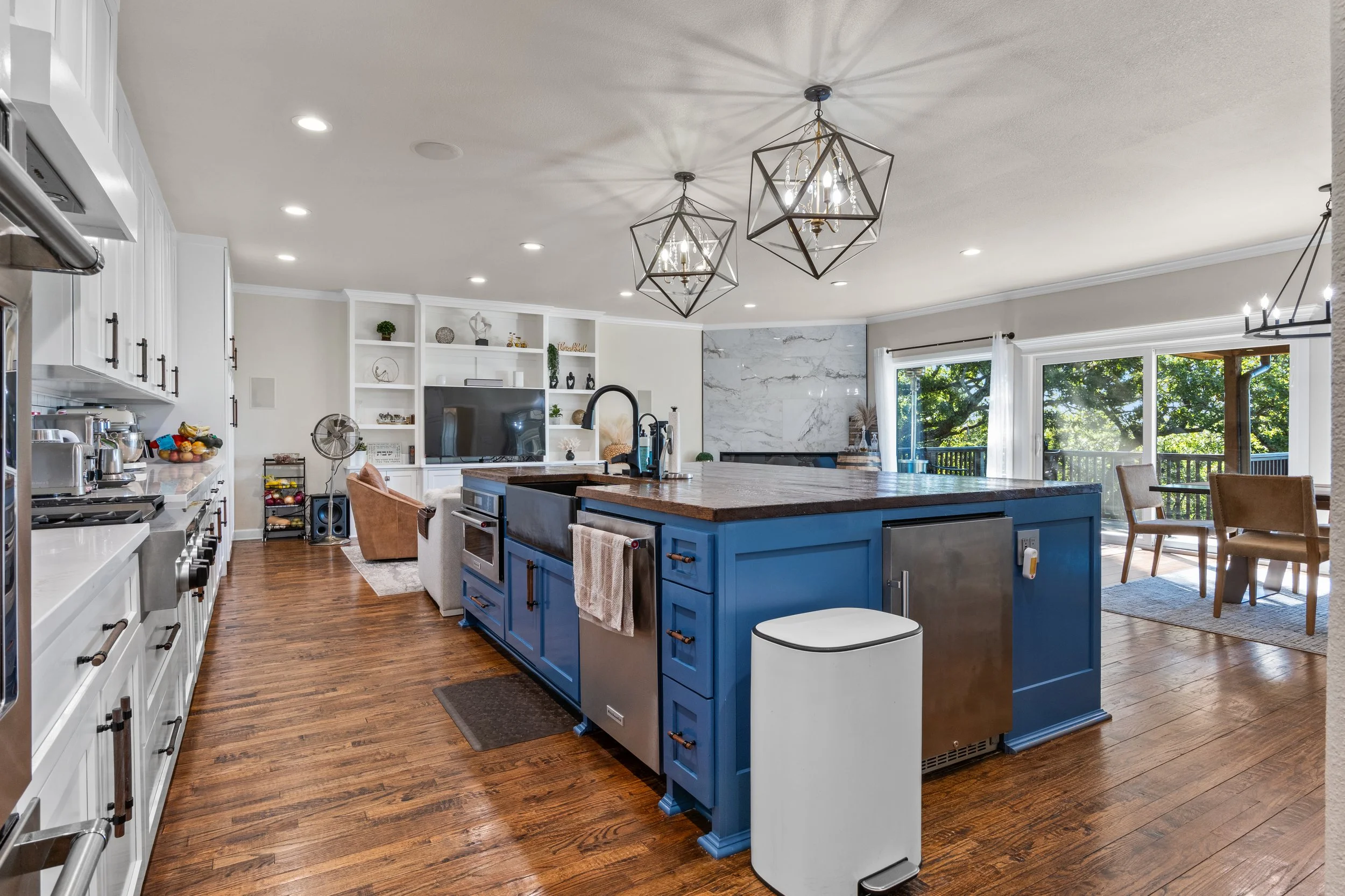 A spacious open-concept kitchen and living room area with hardwood floors, featuring a large blue kitchen island, white cabinetry, and modern dining and living spaces with large windows overlooking a greenery outdoor view.