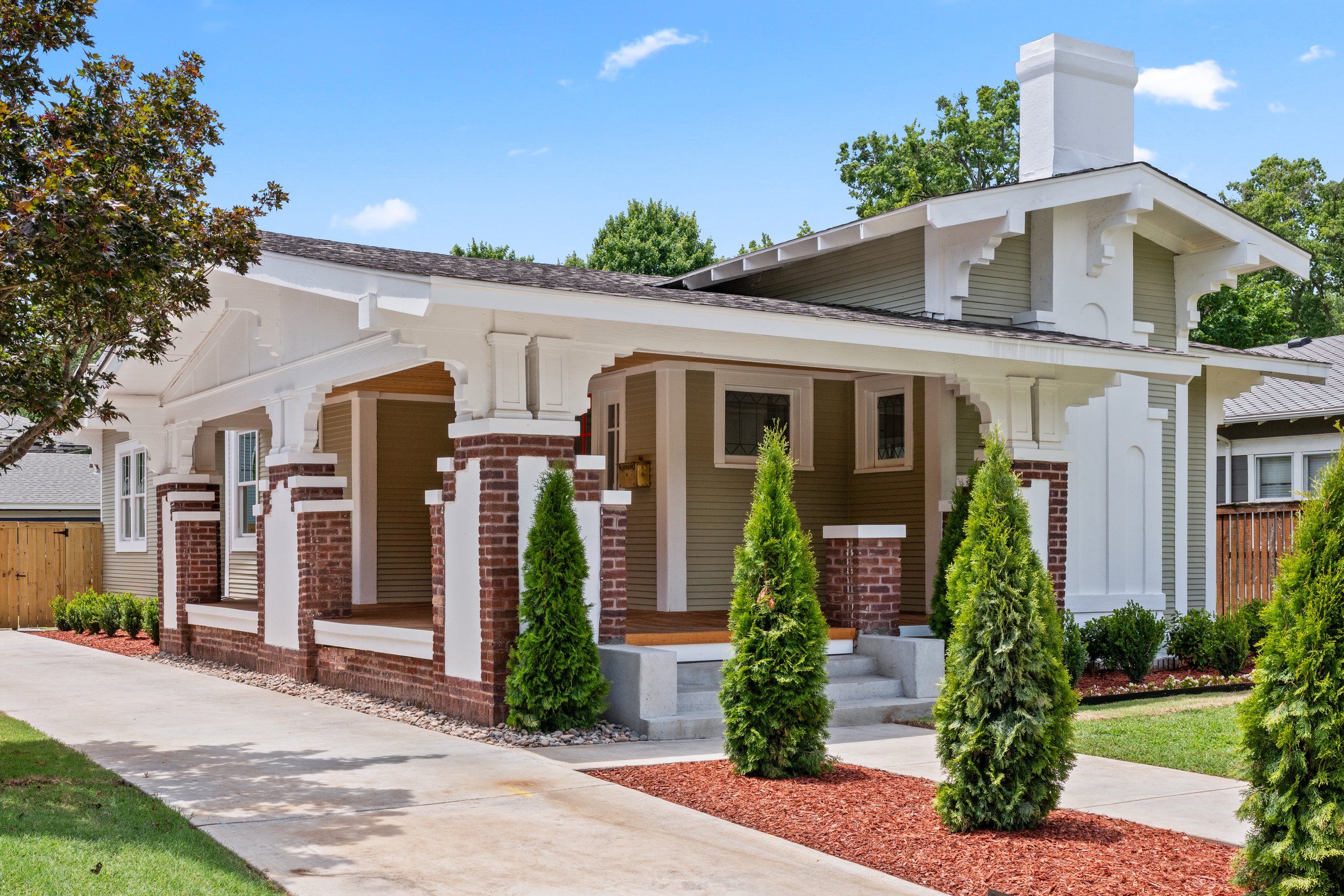 Front view of a house with a porch, brick and white painted wood exterior, surrounded by green trees and shrubs, under a blue sky.