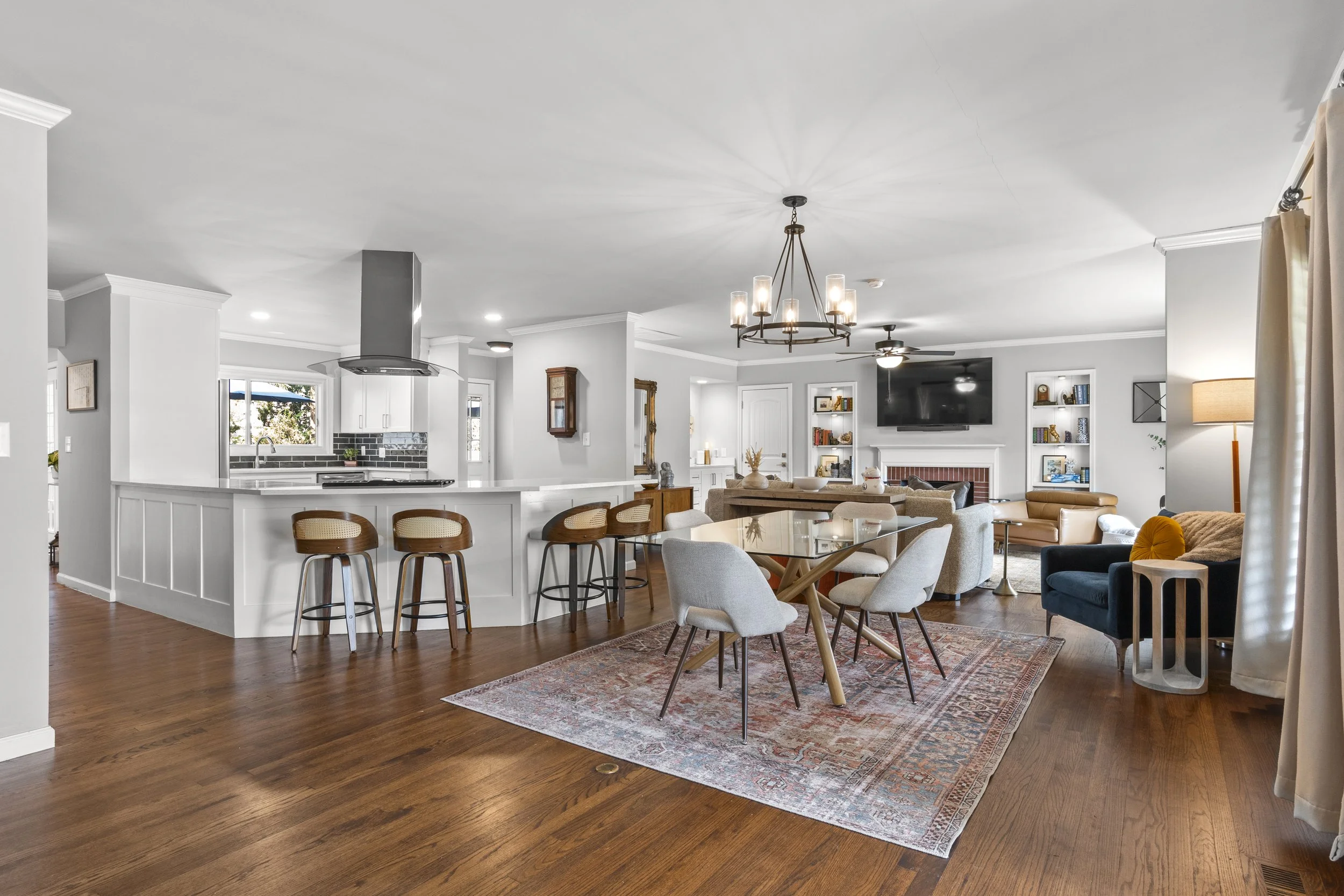 Open concept living space with a kitchen, dining area, and sitting area. Features white walls, hardwood floors, modern furniture, a chandelier, and a large TV above the fireplace.