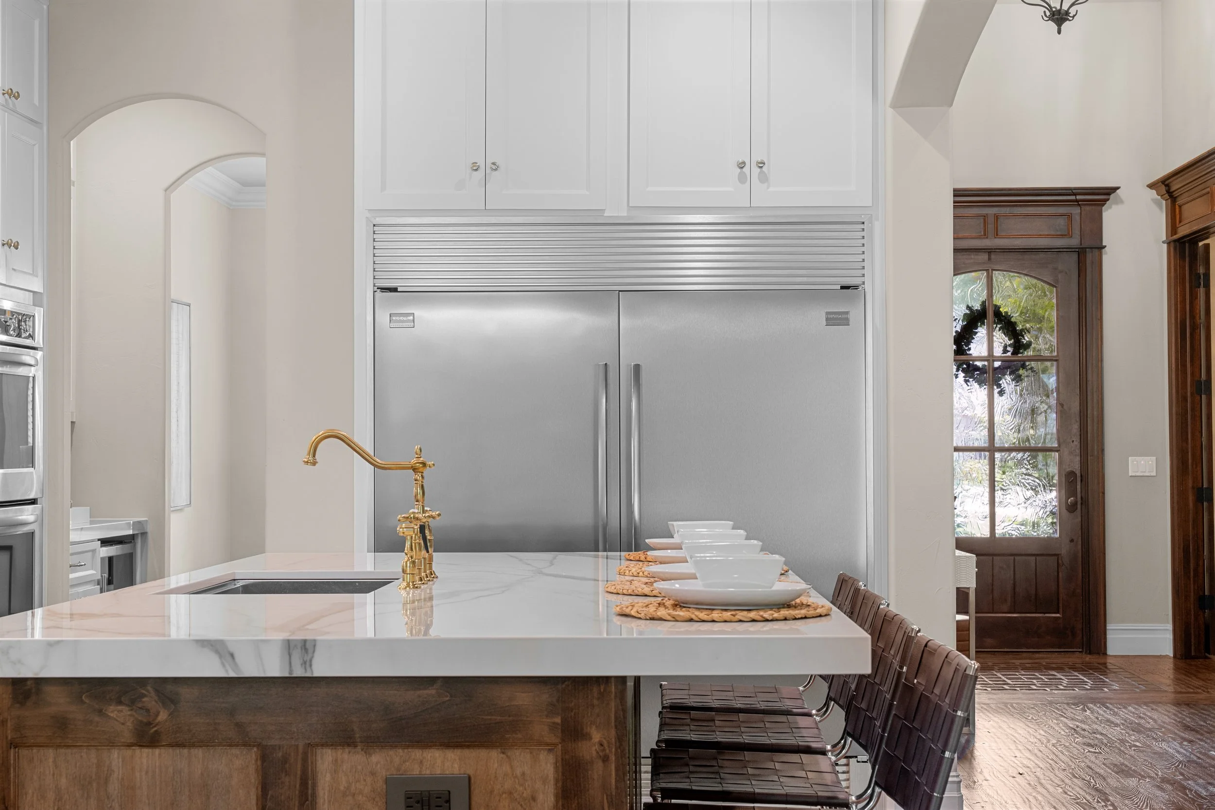 Modern kitchen with white cabinets, stainless steel refrigerators, a marble island with a gold faucet, and four place settings with bowls and plates on woven placemats.