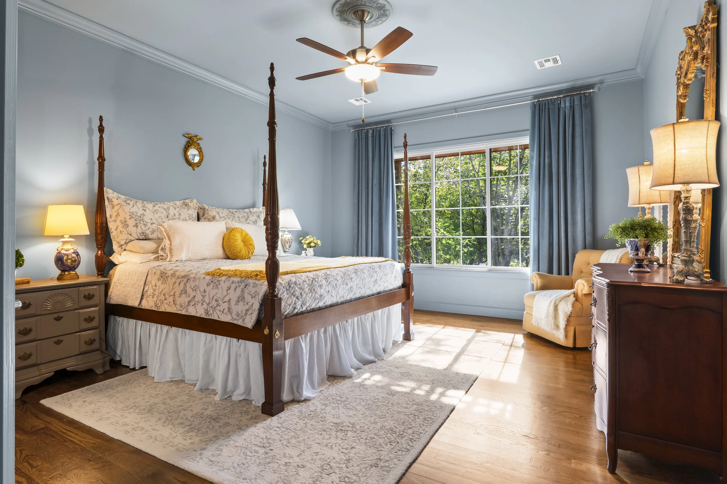 A cozy bedroom with a four-poster bed, a yellow tufted armchair, and a dresser. The room has light blue walls, a large window with blue curtains, and hardwood floors. Decor includes lamps, a small mirror, and a potted plant.