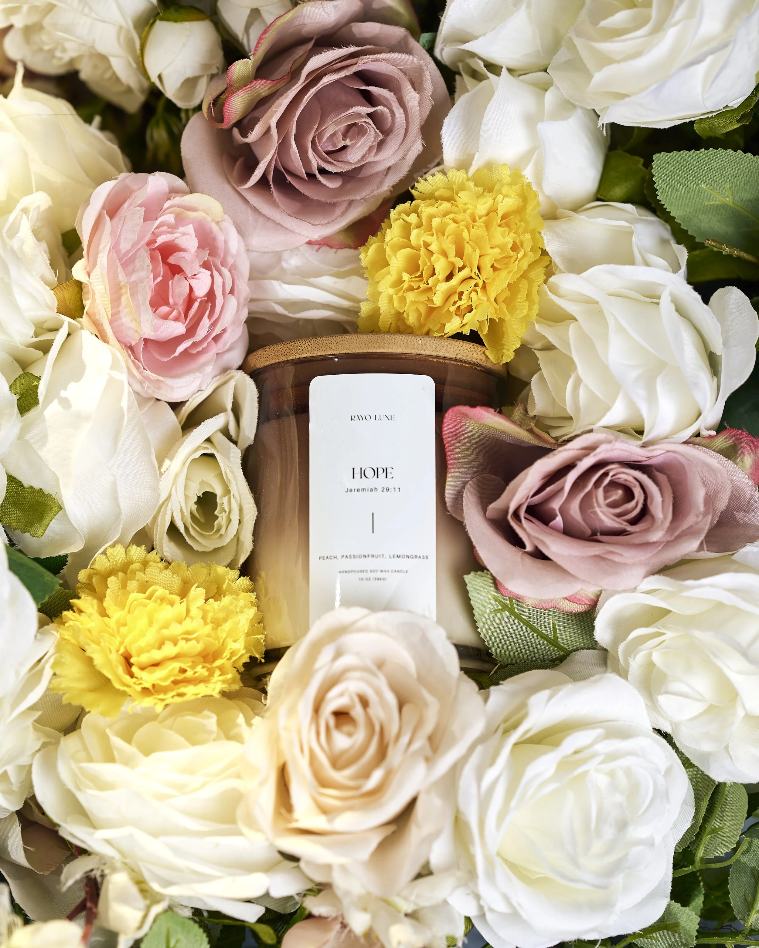 A close-up view of a cluster of artificial flowers, including white roses, pink roses, a yellow marigold, and greenery surrounding a brown candle with a white label.