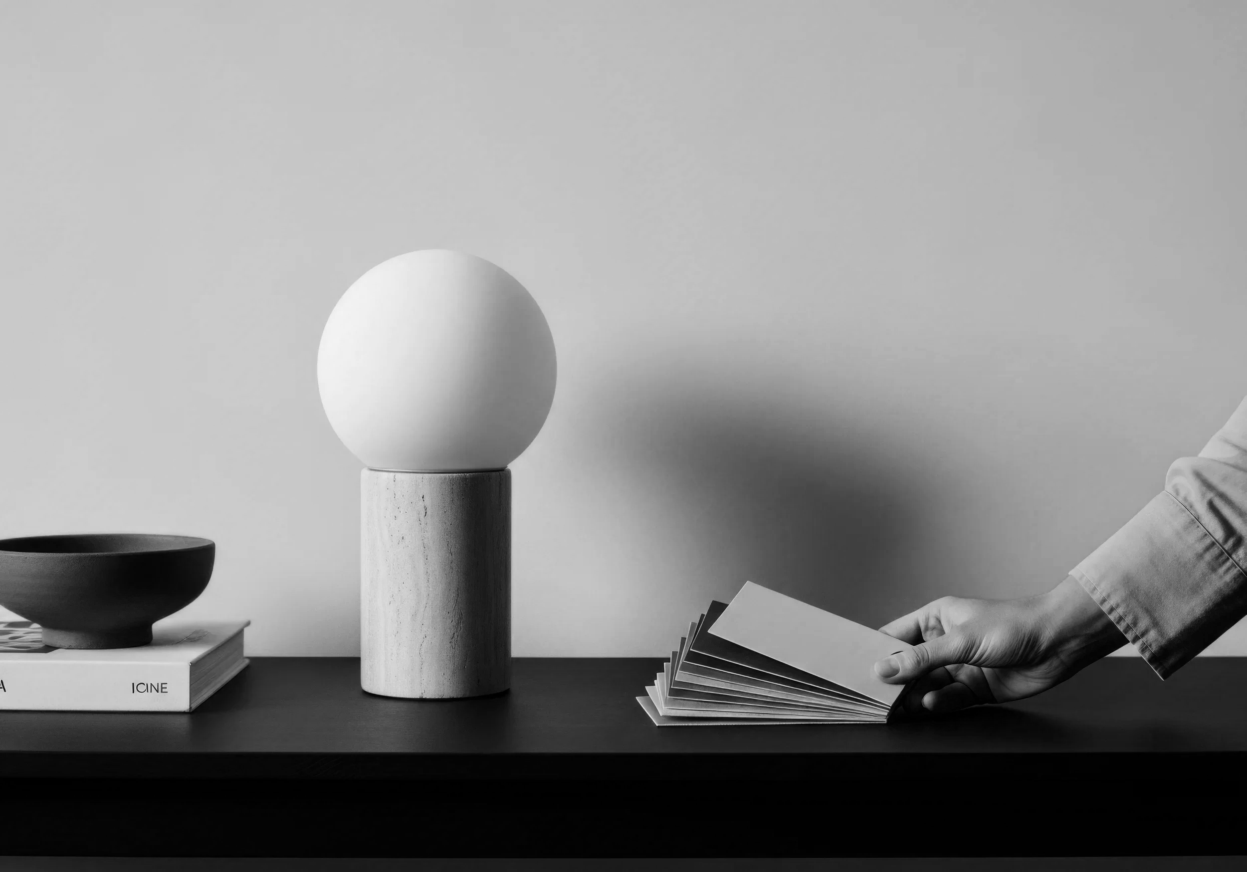 A black and white photograph features a person's hand holding a fan of photographs, a small bowl, a book titled 'ICINE', and a minimalist decorative setup that includes a wooden cylinder with a large egg-shaped object on top, all arranged on a table against a plain background.