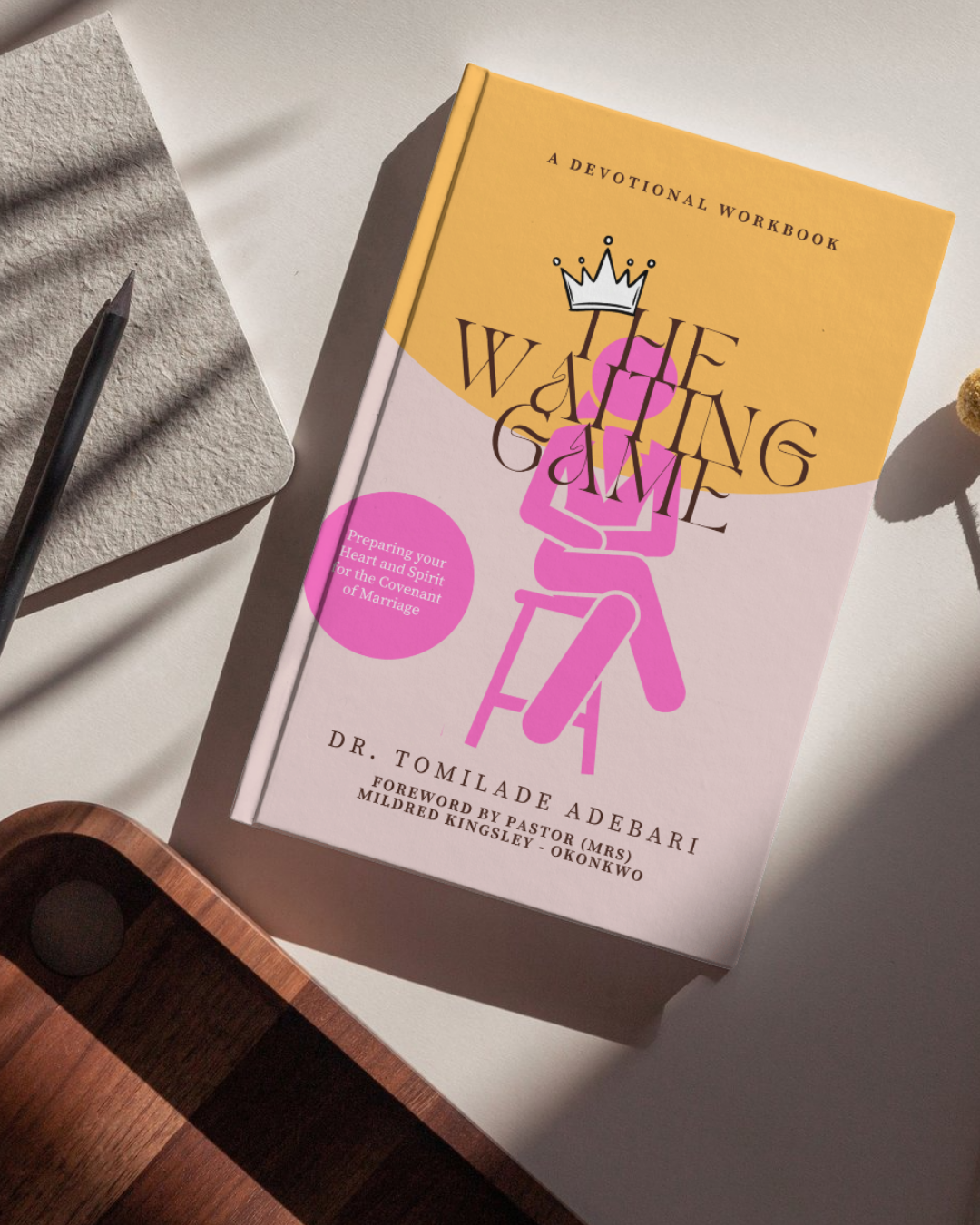 Book titled 'The Winning Game' with a pink outline of a woman sitting and a crown on her head, on a white surface with a notepad, pencil, and a wooden object nearby.