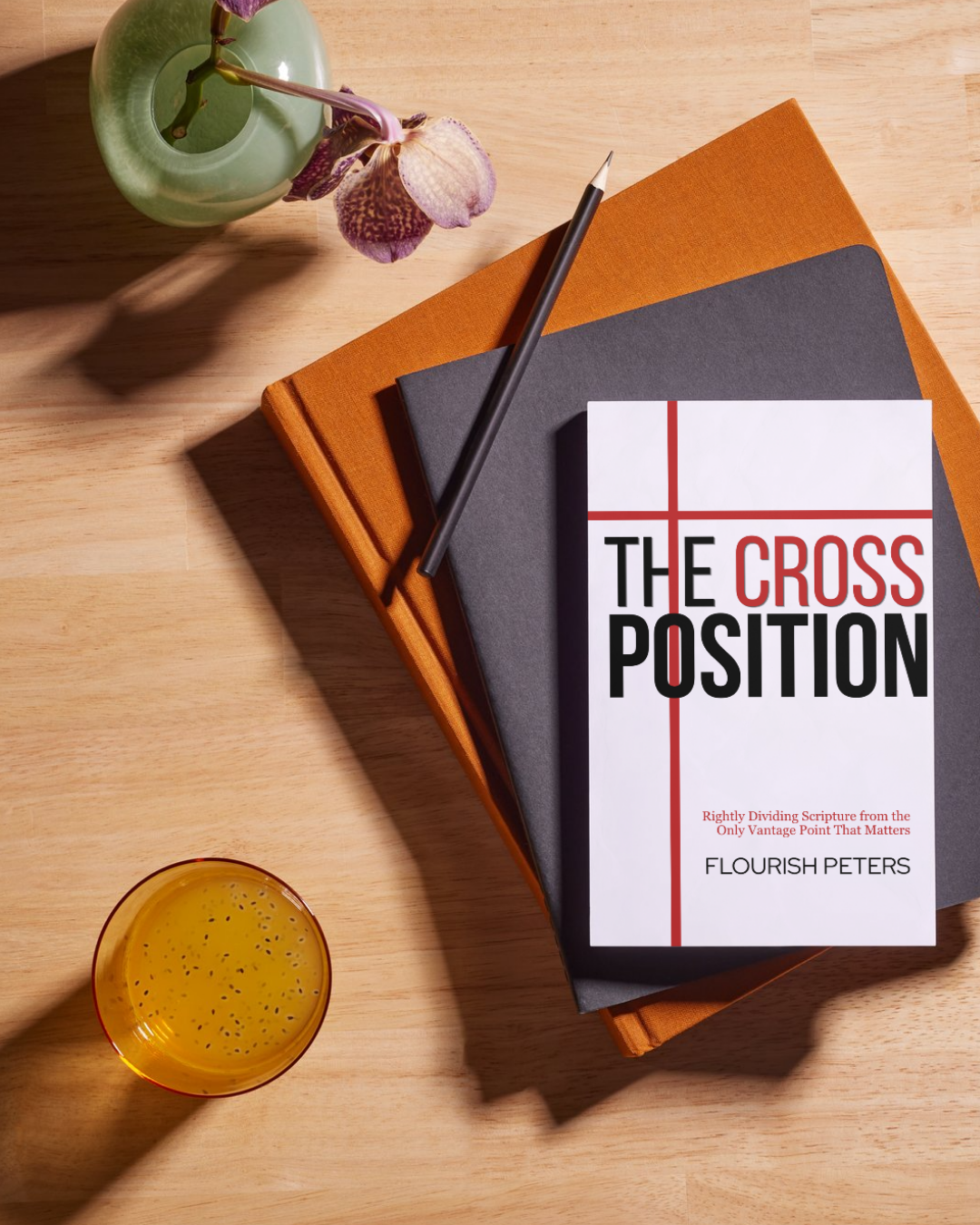 A wooden table holding a green vase with purple orchids, a stack of two hardcover books (orange and gray), a white book titled 'The Cross Position' by Flourish Peters, a black pen, and a glass of orange juice with tiny black seeds.