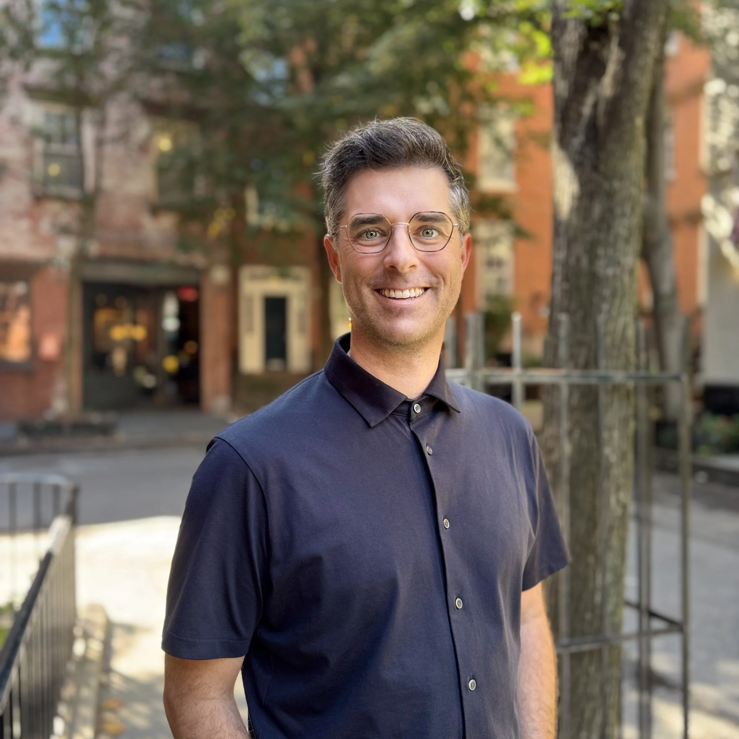 Our pastor, smiling and standing on a tree-lined New York City block, with short dark hair and glasses.