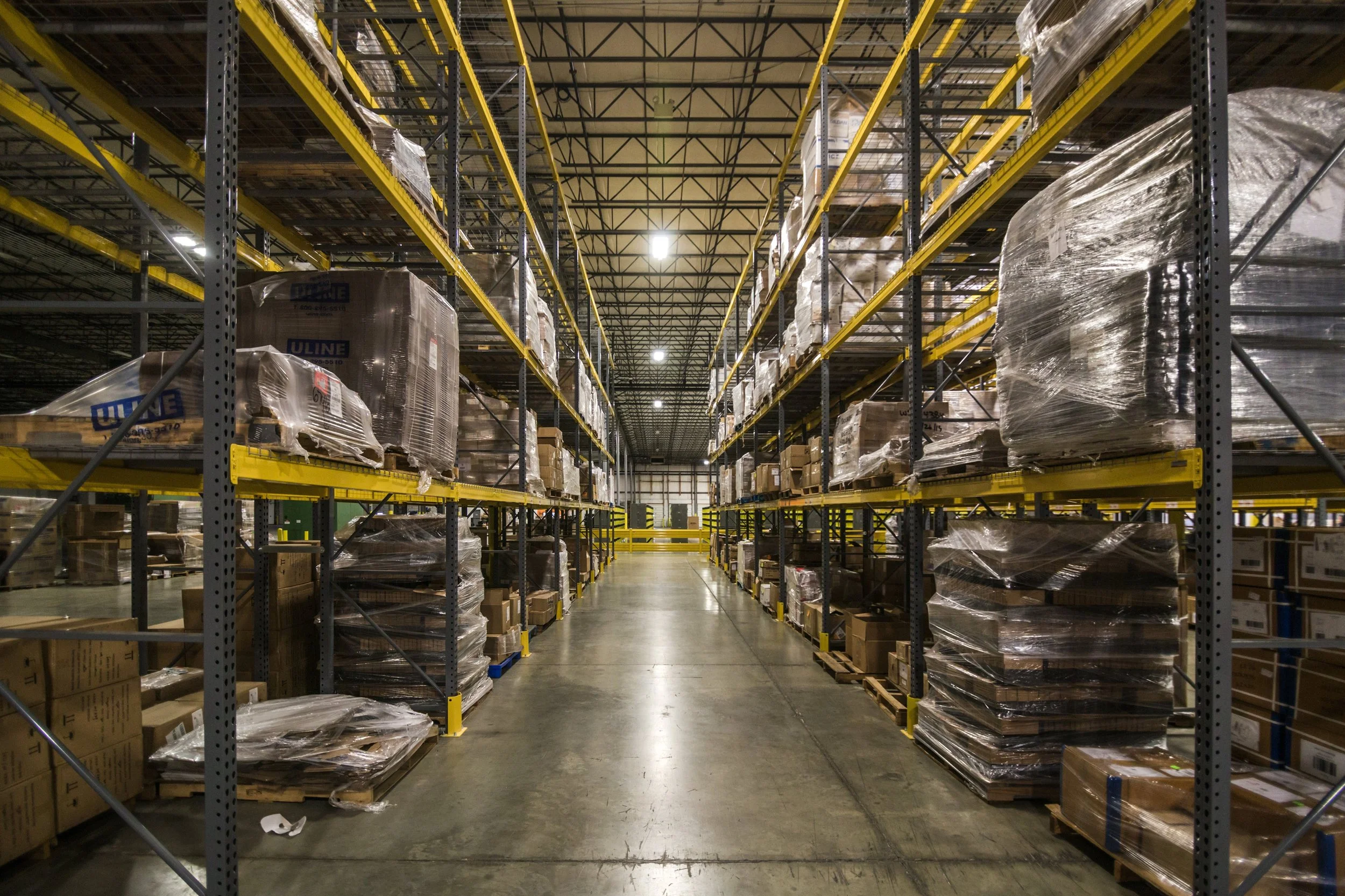 Empty warehouse aisle with tall metal shelves stocked with cardboard boxes and pallets wrapped in plastic.