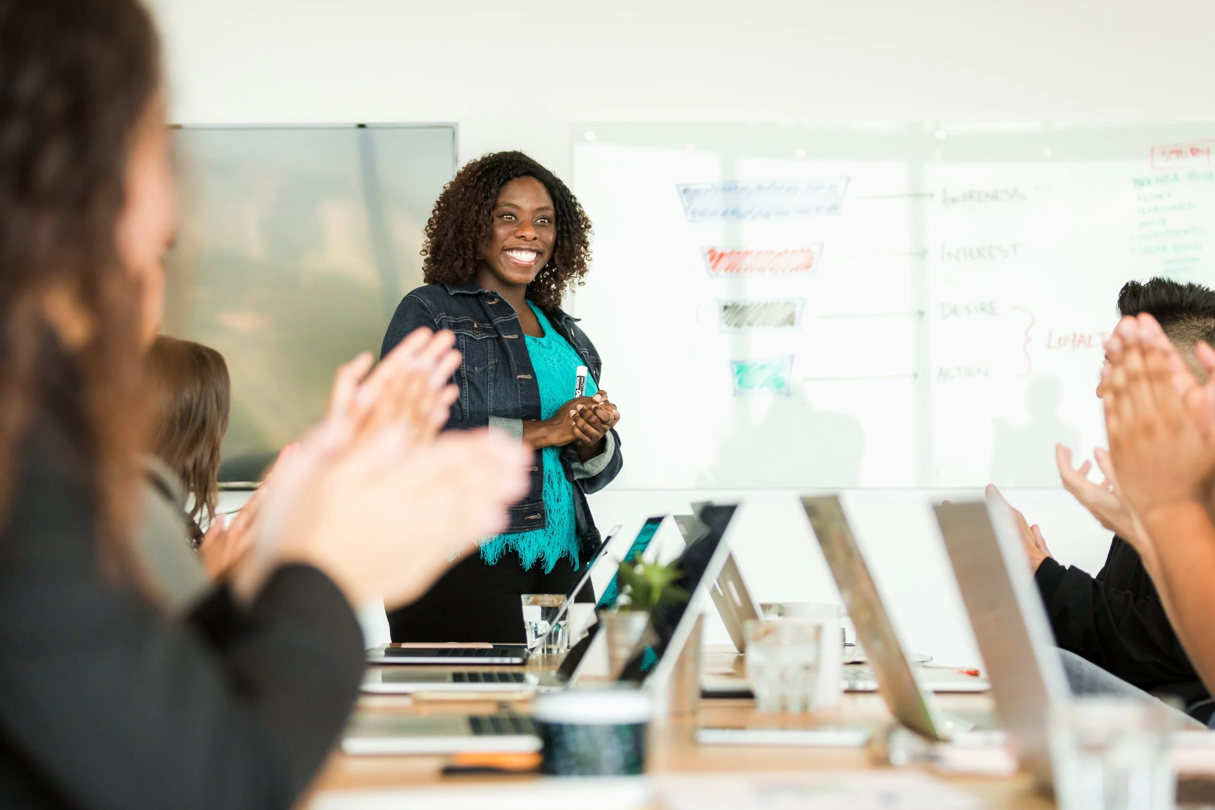 A woman standing and smiling in front of a classroom or meeting room whiteboard with notes; people seated at a table with laptops, clapping and engaging.