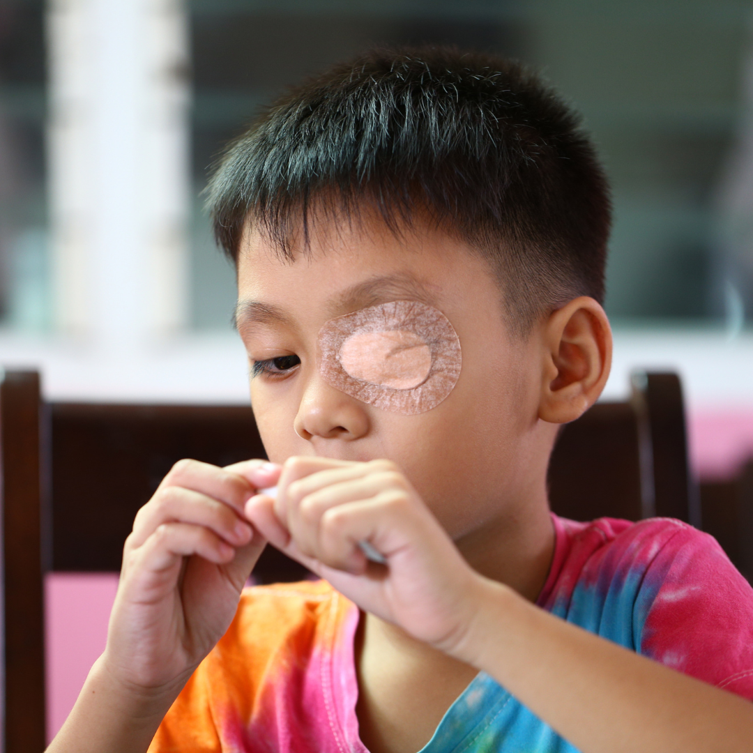 A young boy with a patch over his left eye sitting at a table, wearing a tie-dye shirt, and appearing to be focusing on something in his hands.