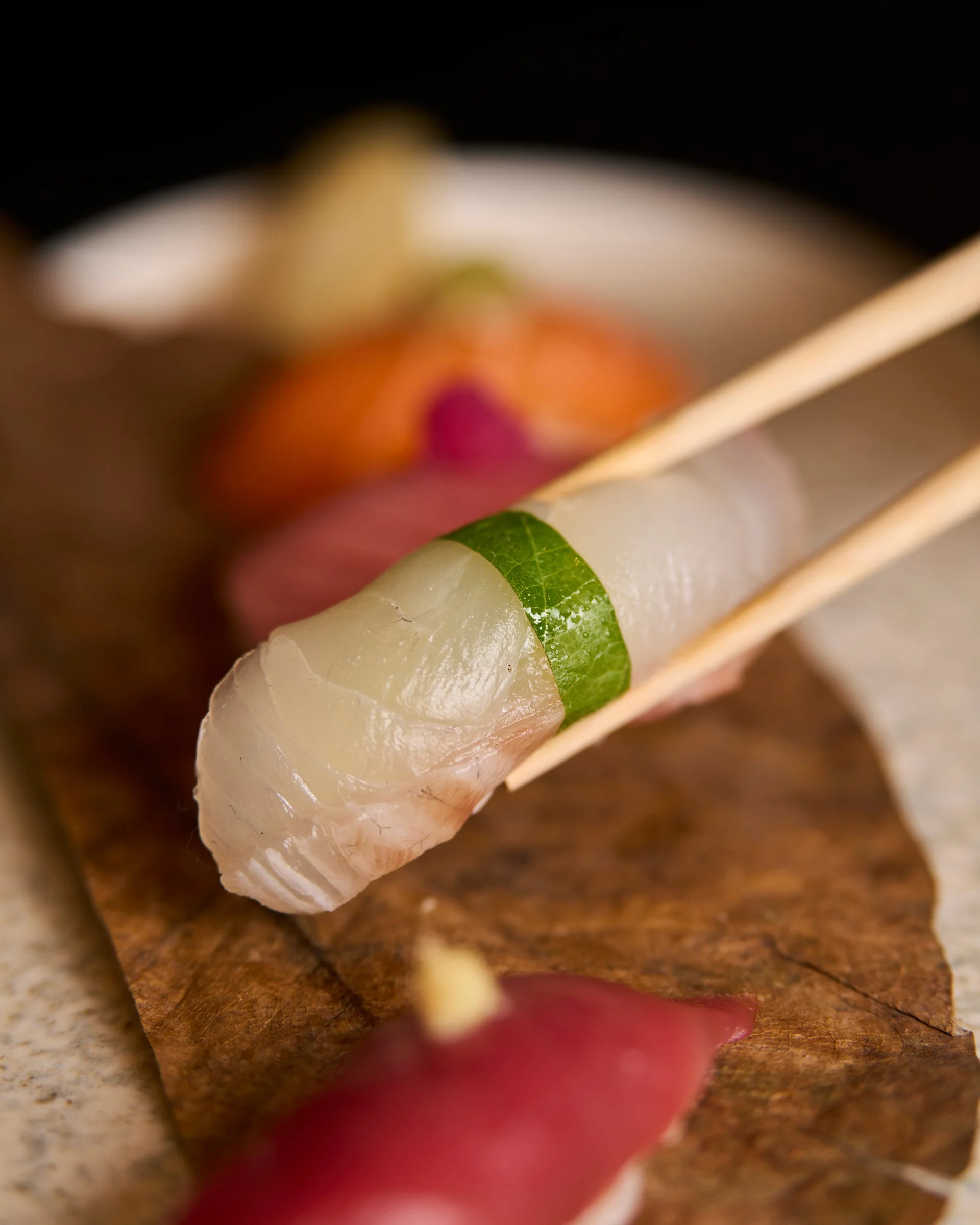 Close-up of a piece of sashimi, possibly white fish, held with chopsticks. In the background, various other sashimi pieces, including tuna and salmon, are on a wooden surface.