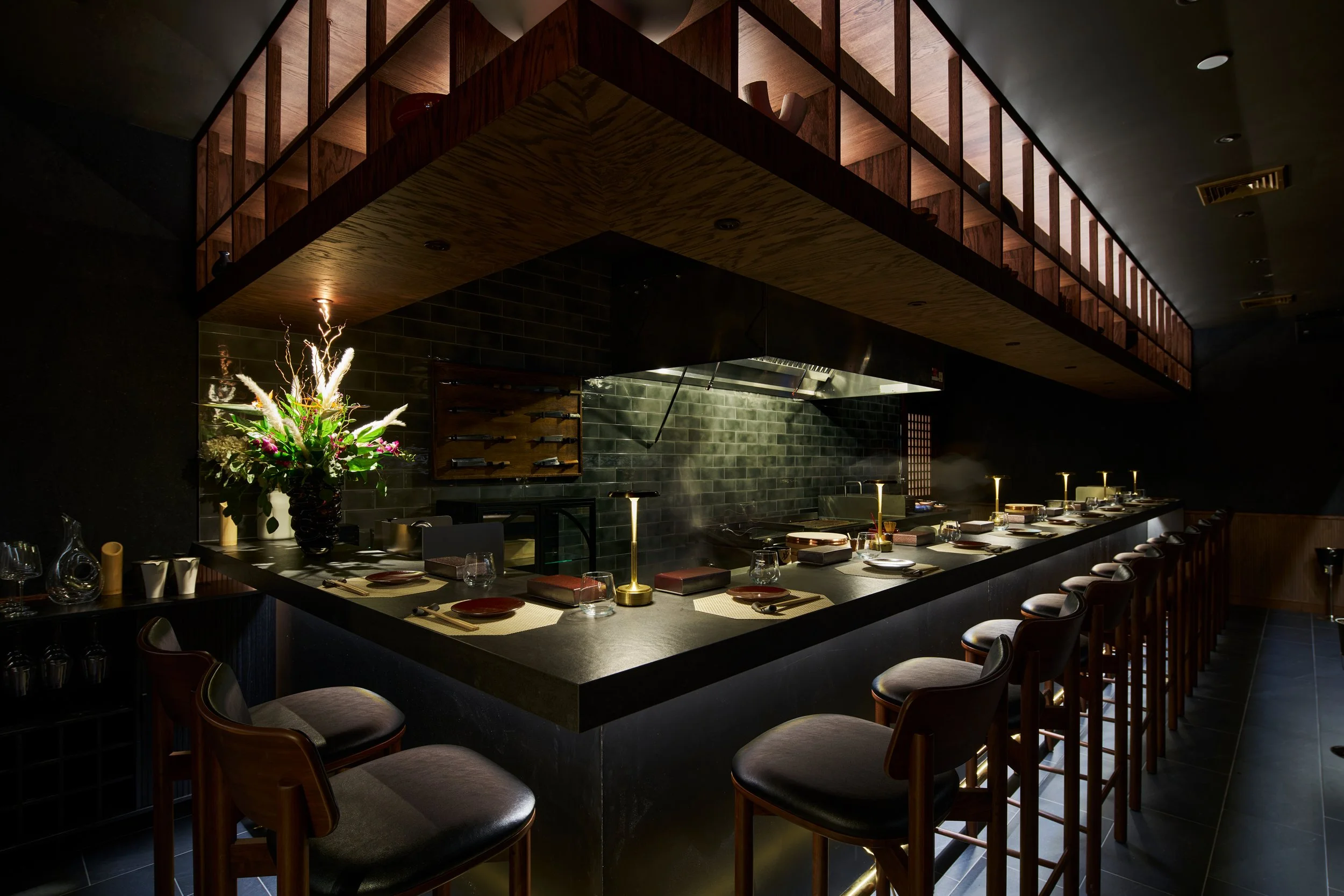A modern, dimly lit restaurant with a black countertop bar and seating area. The bar is decorated with a large flower arrangement, wine glasses, and plates. The background features a black tiled wall, a wooden rack with knives, and sizzling cooking a