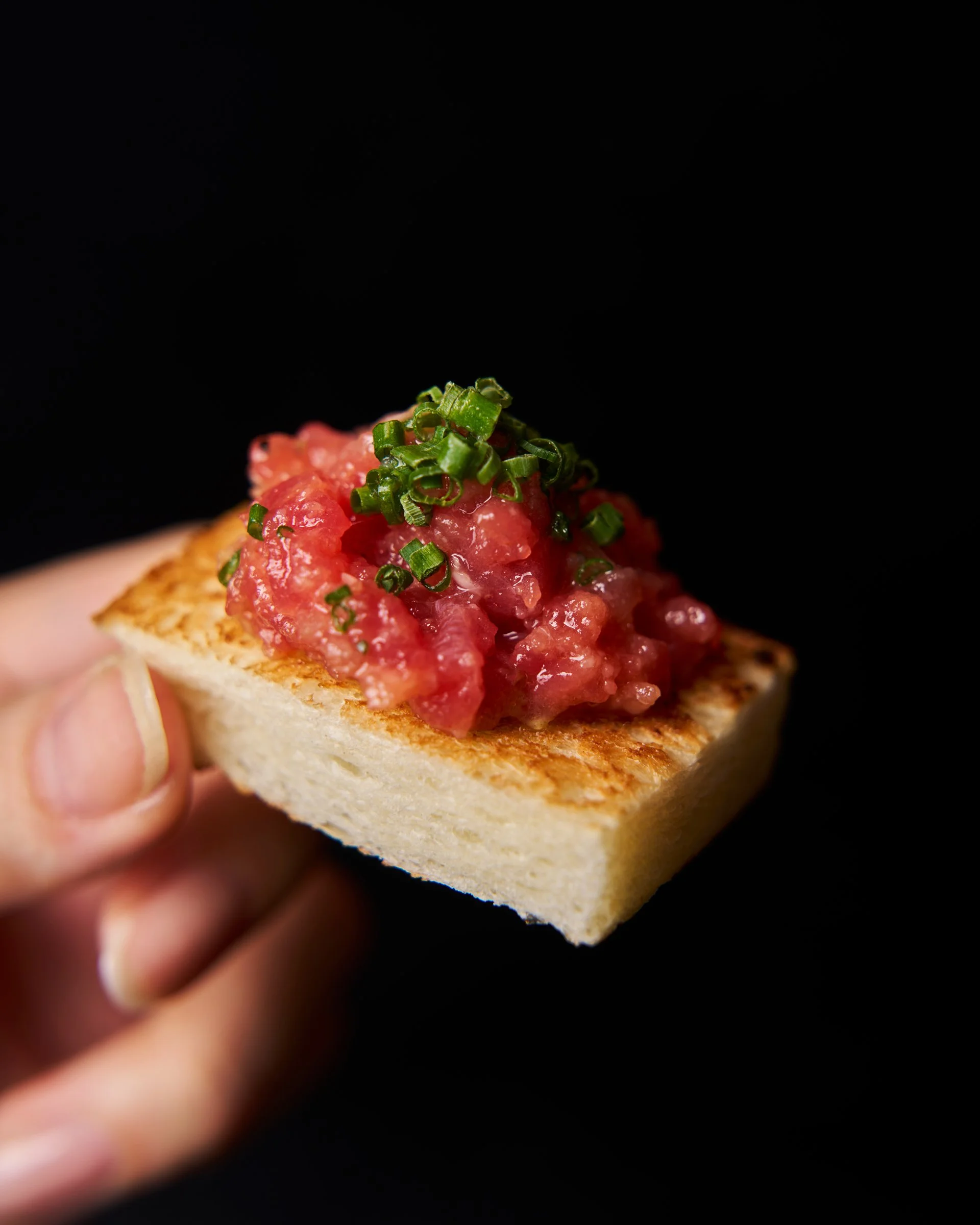 A hand holding a toasted piece of bread topped with chopped tomatoes and chopped green herbs against a black background.