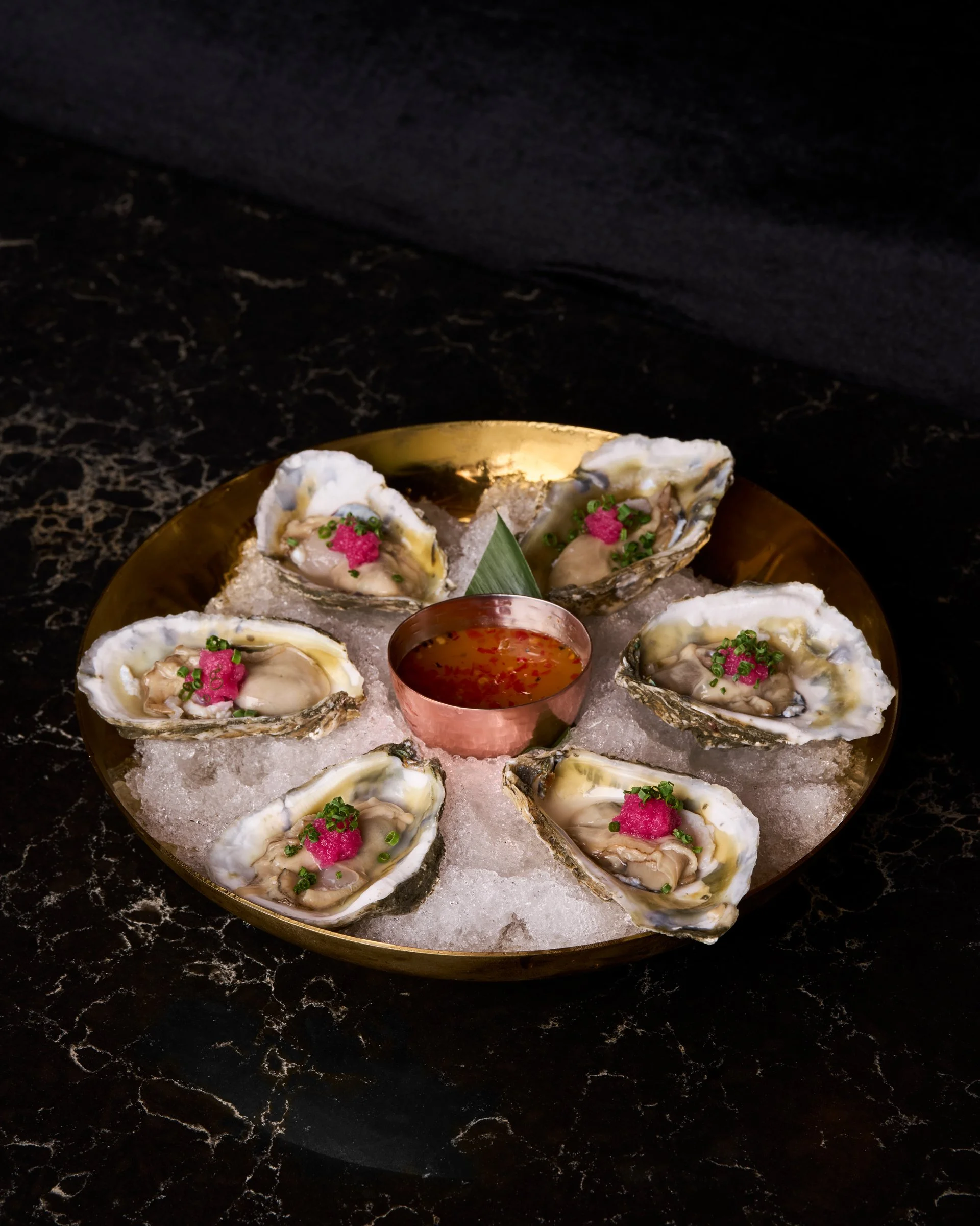 A round golden plate with seven oysters on ice, garnished with pink roe and chives, with a small copper bowl of sauce in the center on a dark marble surface.