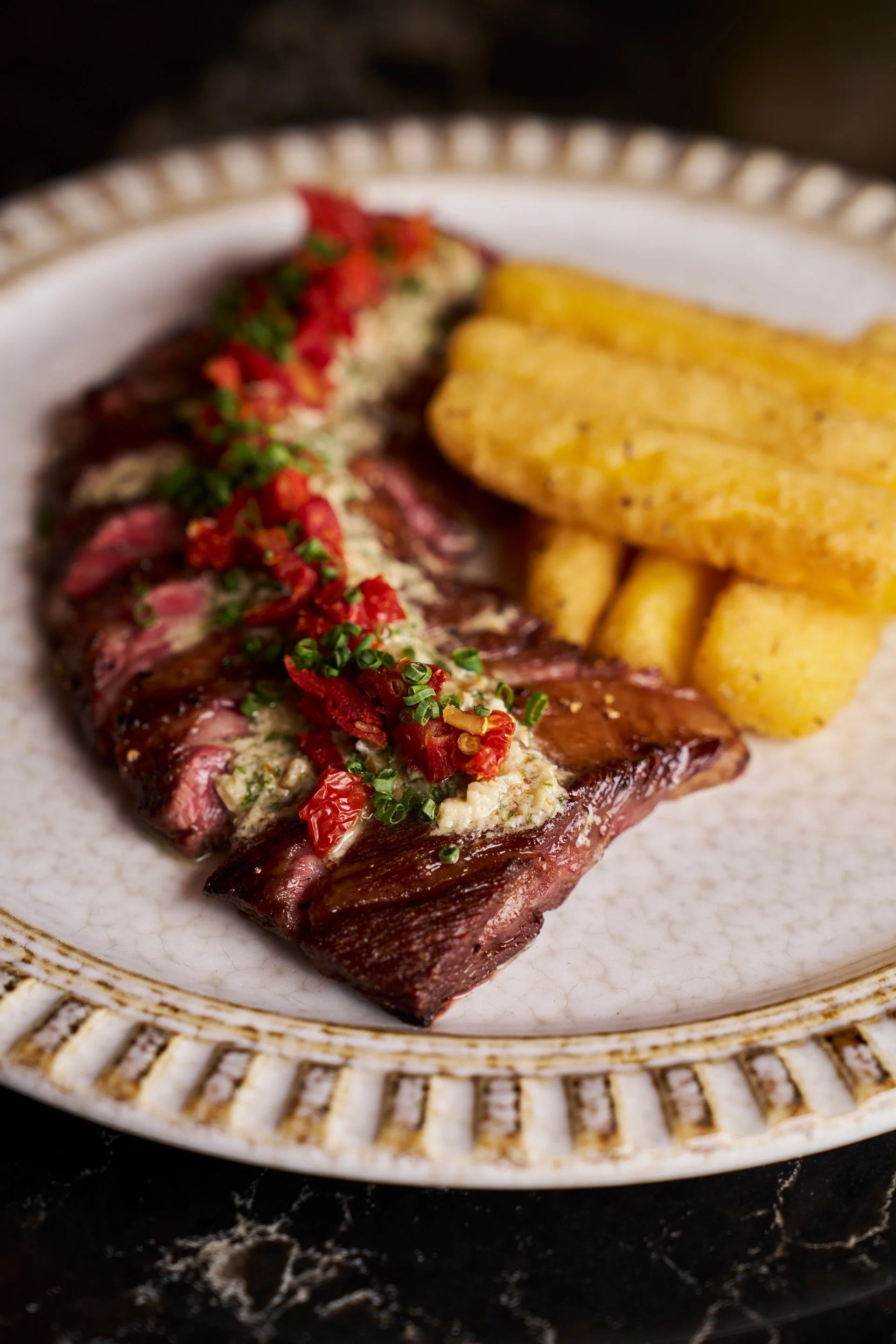 A plate of sliced steak topped with chopped chives and red peppers, served with fried plantains.