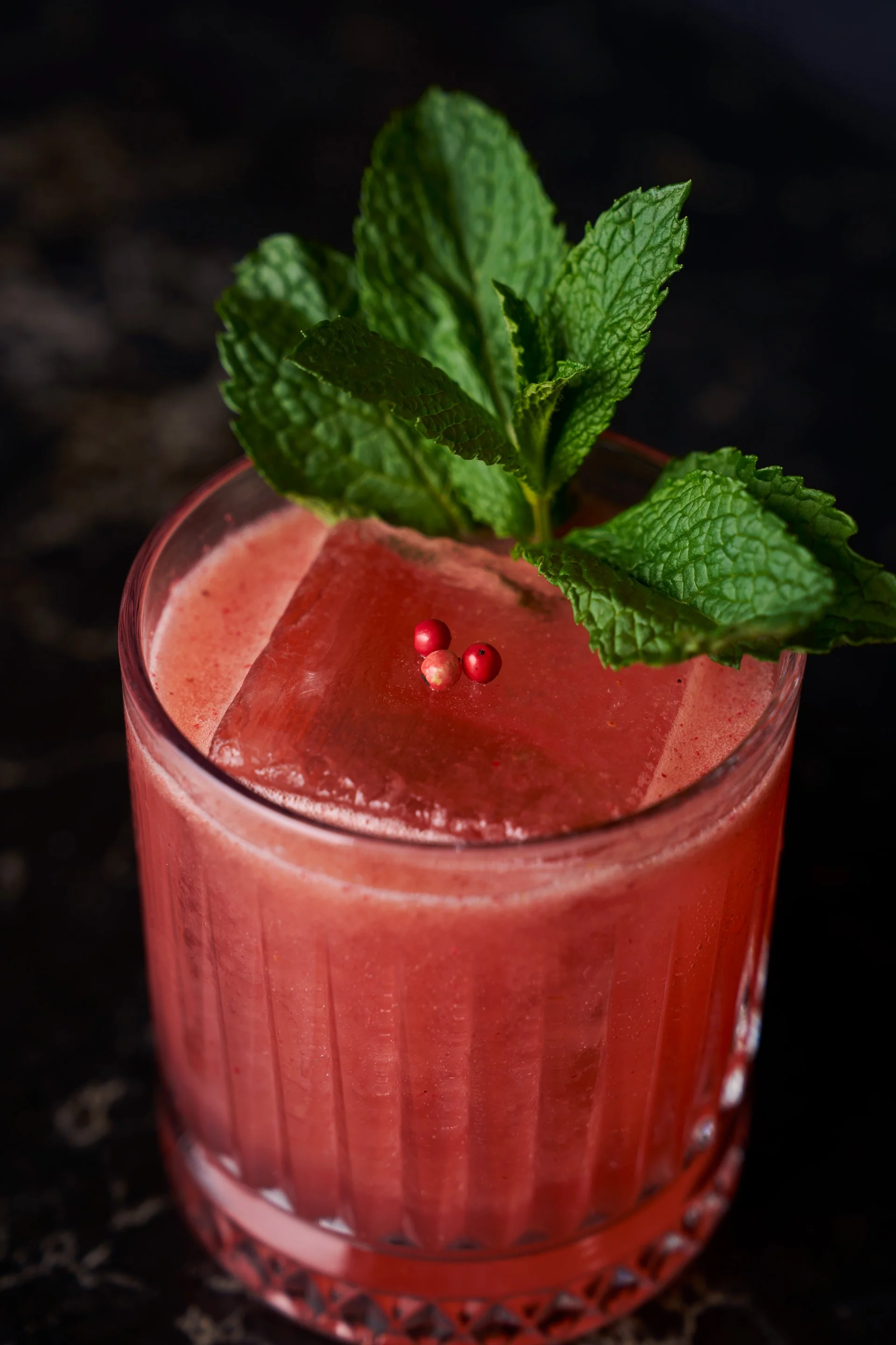 A pink cocktail garnished with fresh mint leaves, red peppercorns, and a large ice cube in a textured glass on a dark background.