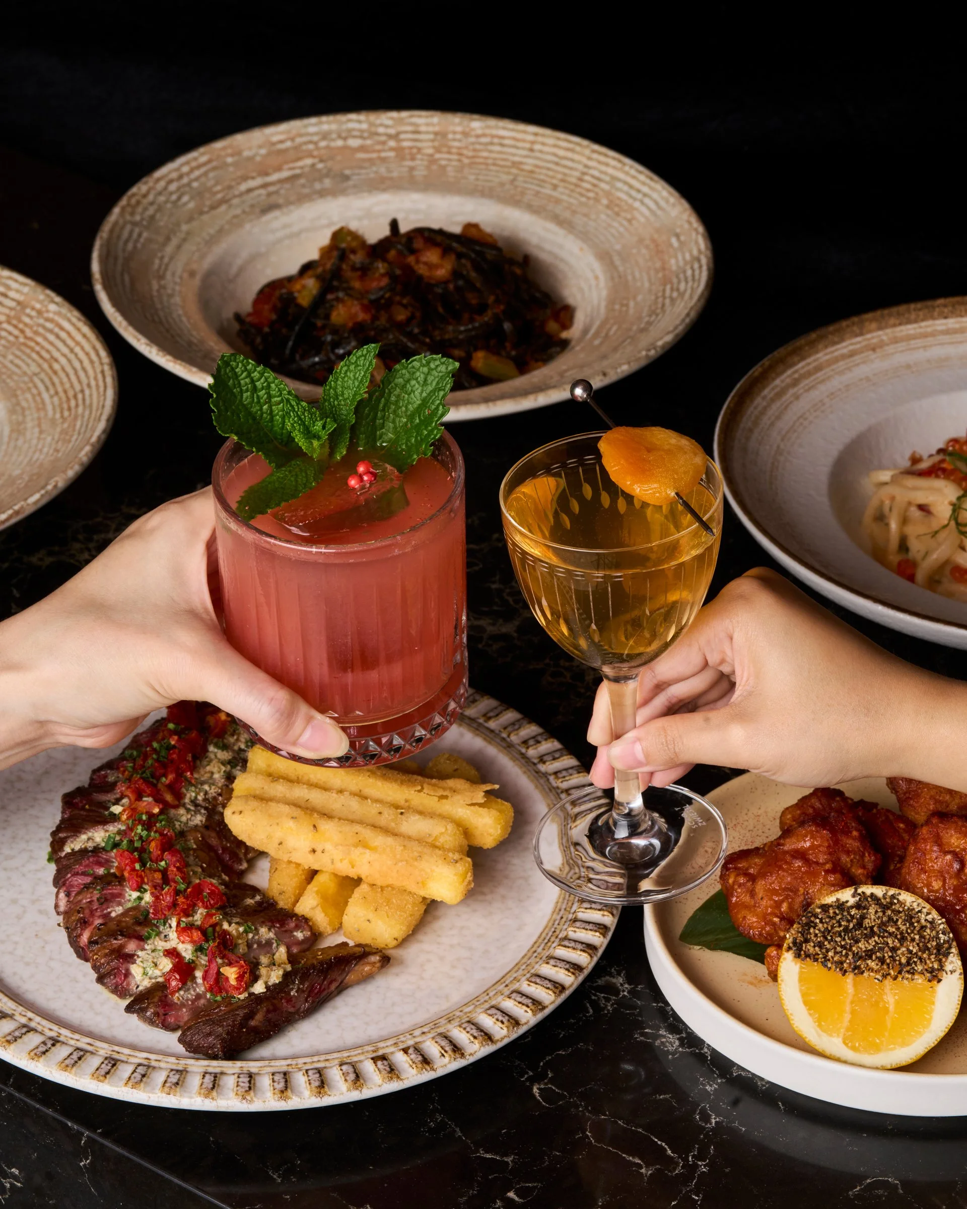 Two people toasting with cocktails surrounded by various plated dishes, including fried food, steak, pasta, and spicy chicken, on a black marble table.