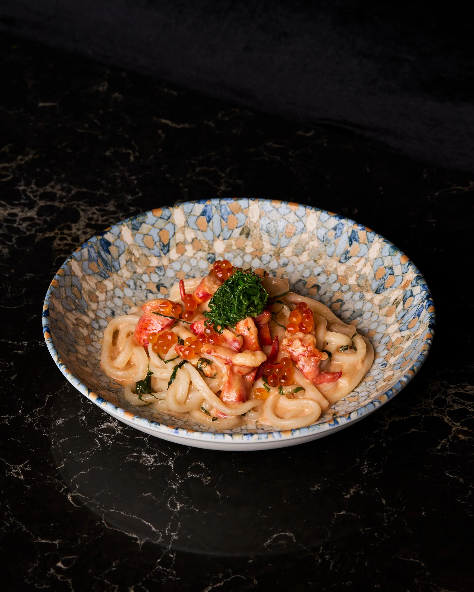 A bowl of creamy pasta topped with red seafood, green herbs, and orange fish roe on a black marble surface.