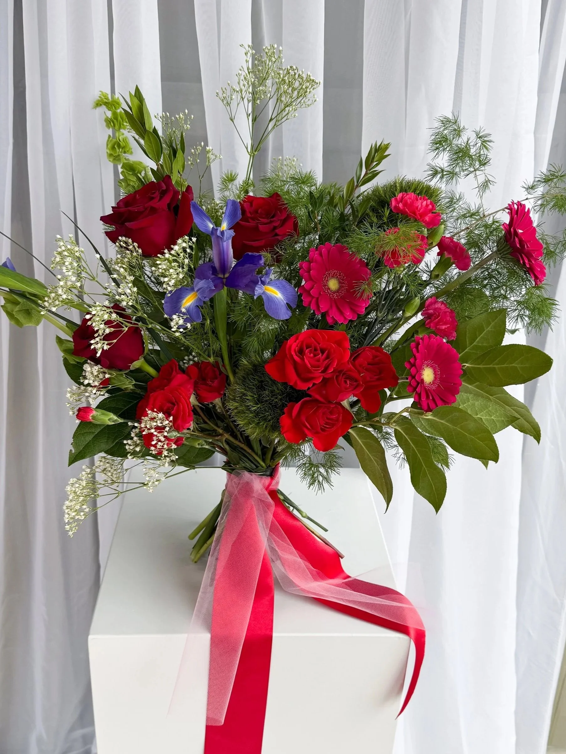 A colorful bouquet of red roses, pink gerbera daisies, purple irises, and white baby's breath flowers wrapped with a pink and red ribbon, placed on a white surface with a sheer white curtain background.