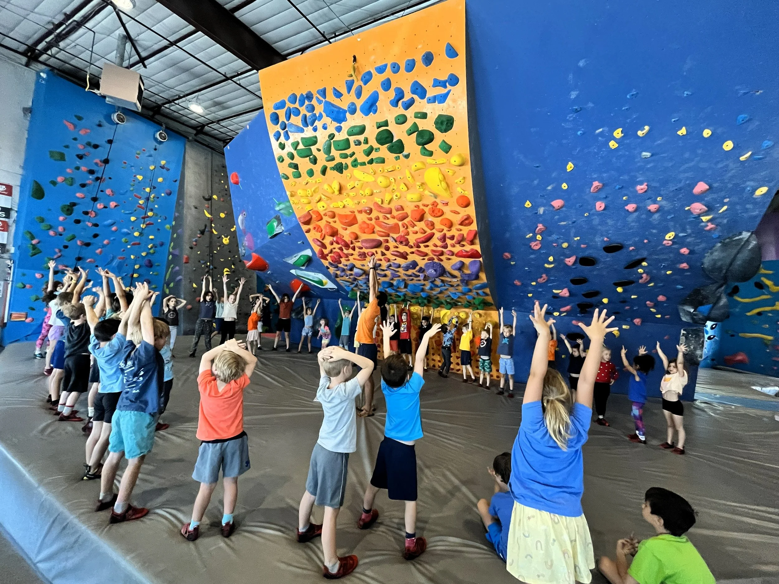 Large group of kids stretching with arms raised beneath tall rope climbing walls during a homeschool climbing club