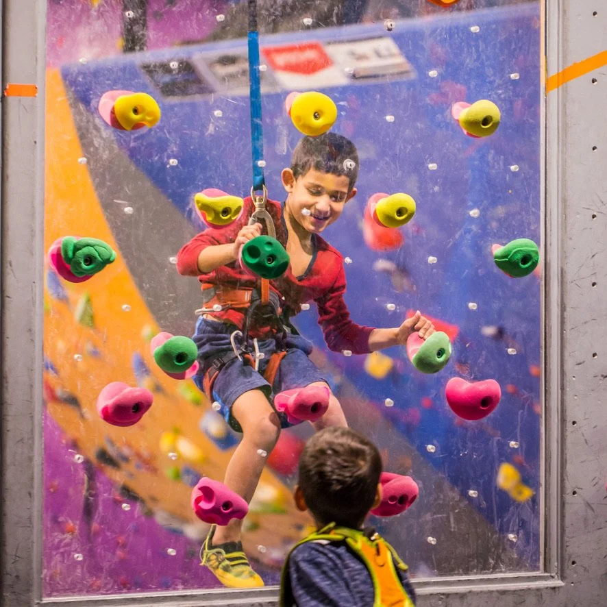 Smiling boy top rope climbing during open gym at ABC Kids Climbing in Boulder while a friend watches