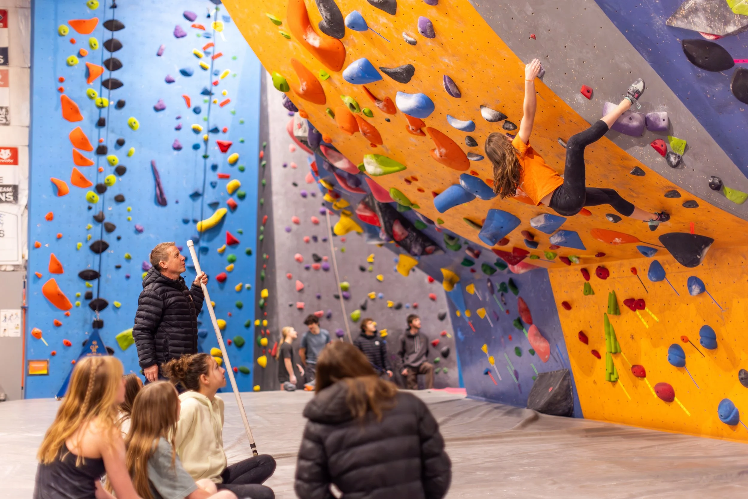 Young climbers watching a teammate on the tall orange wall at ABC Kids Climbing, a climbing gym built for kids in Boulder