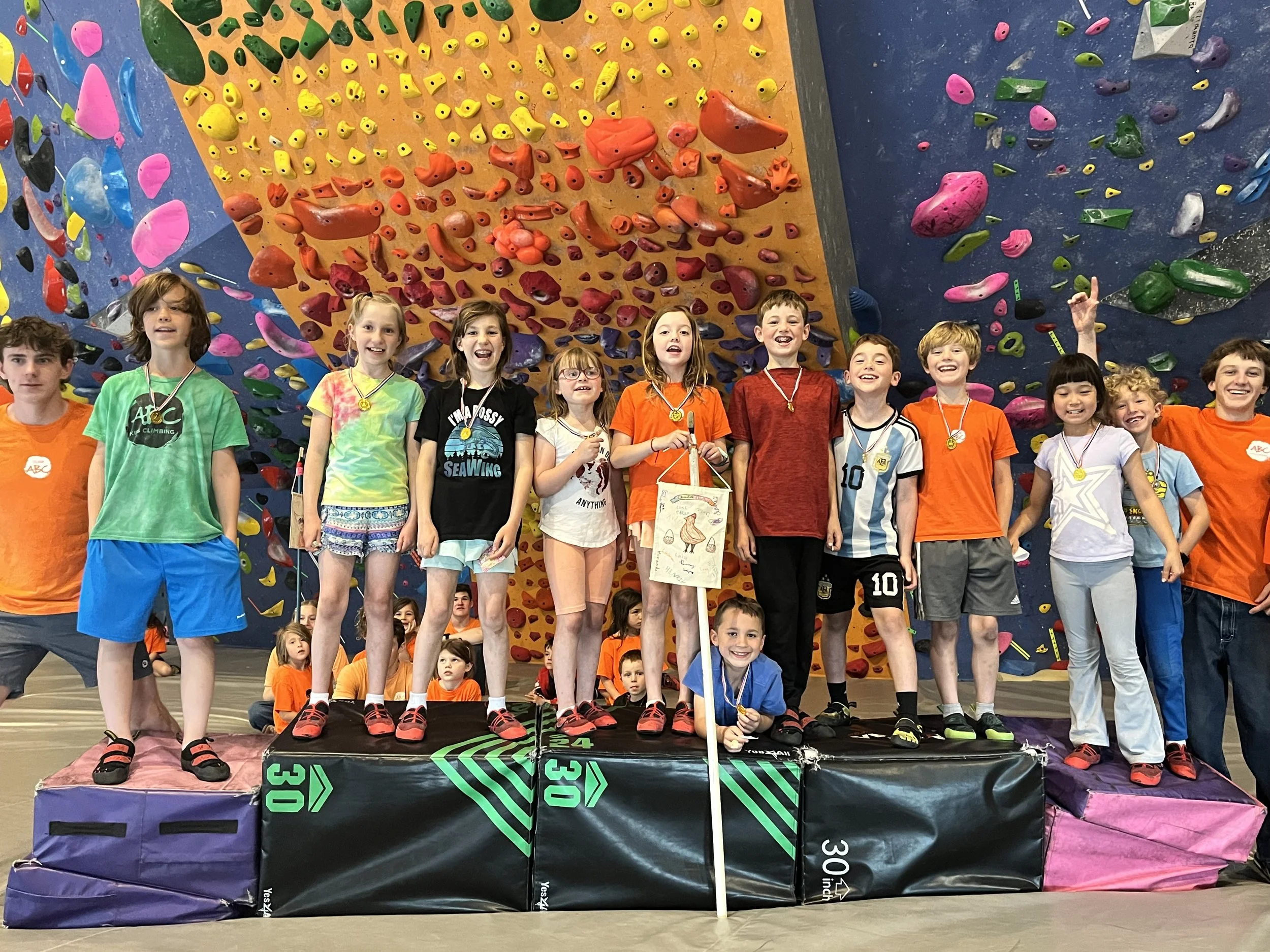 Group of smiling kids posing on a podium in front of the bouldering wall at ABC indoor summer climbing camp in Boulder